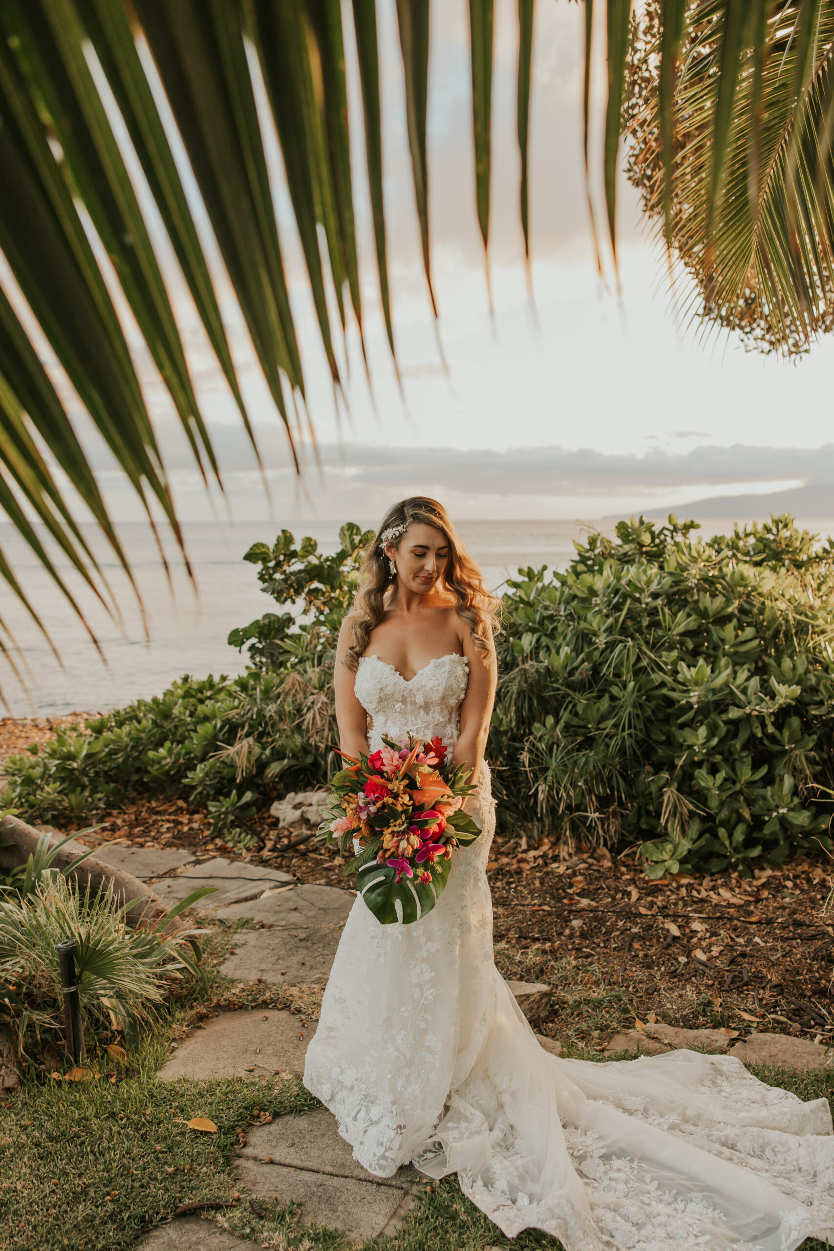 Bride stands beneath tropical foliage, holding her bouquet and glowing in the golden light near the shore at a serene Maui wedding venue.