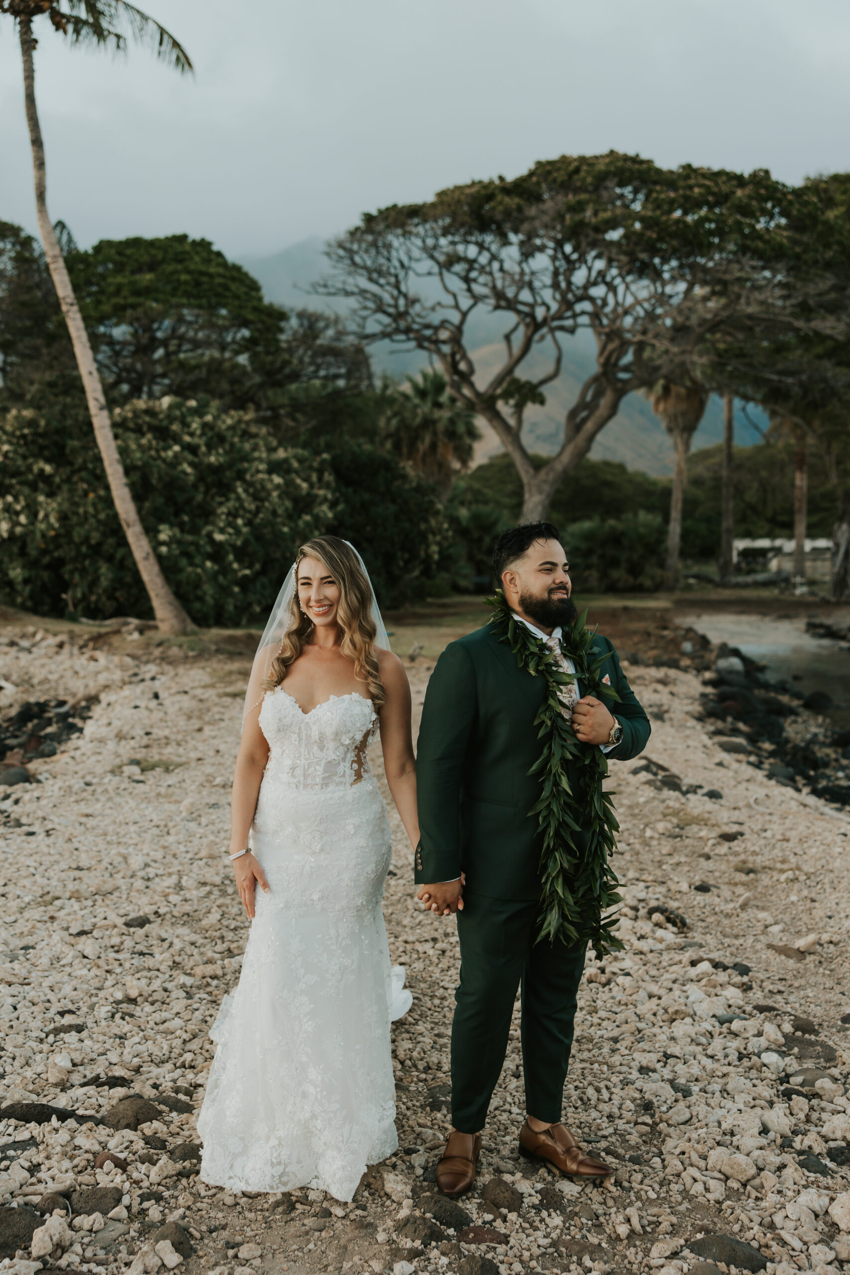 Bride and groom holding hands on a rocky shore with lush trees and misty mountains behind them at a stunning Maui wedding venue.