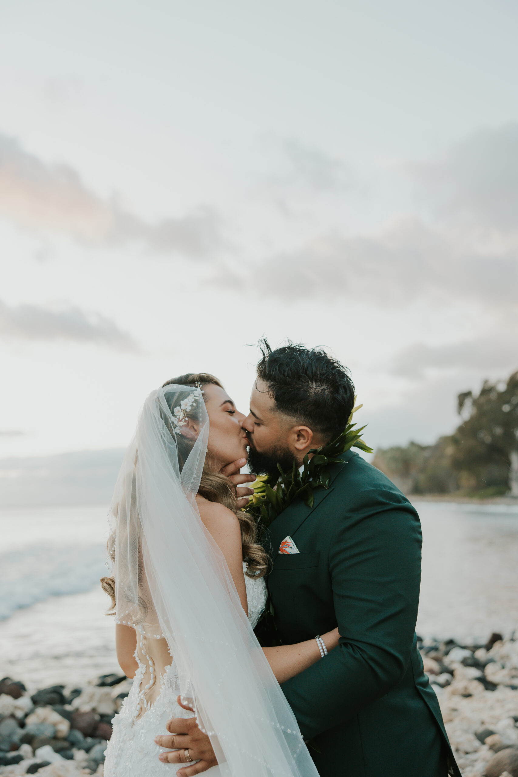 Sweet kiss between the bride and groom on a rocky beach, their silhouettes framed by soft pastel skies and ocean waves.
