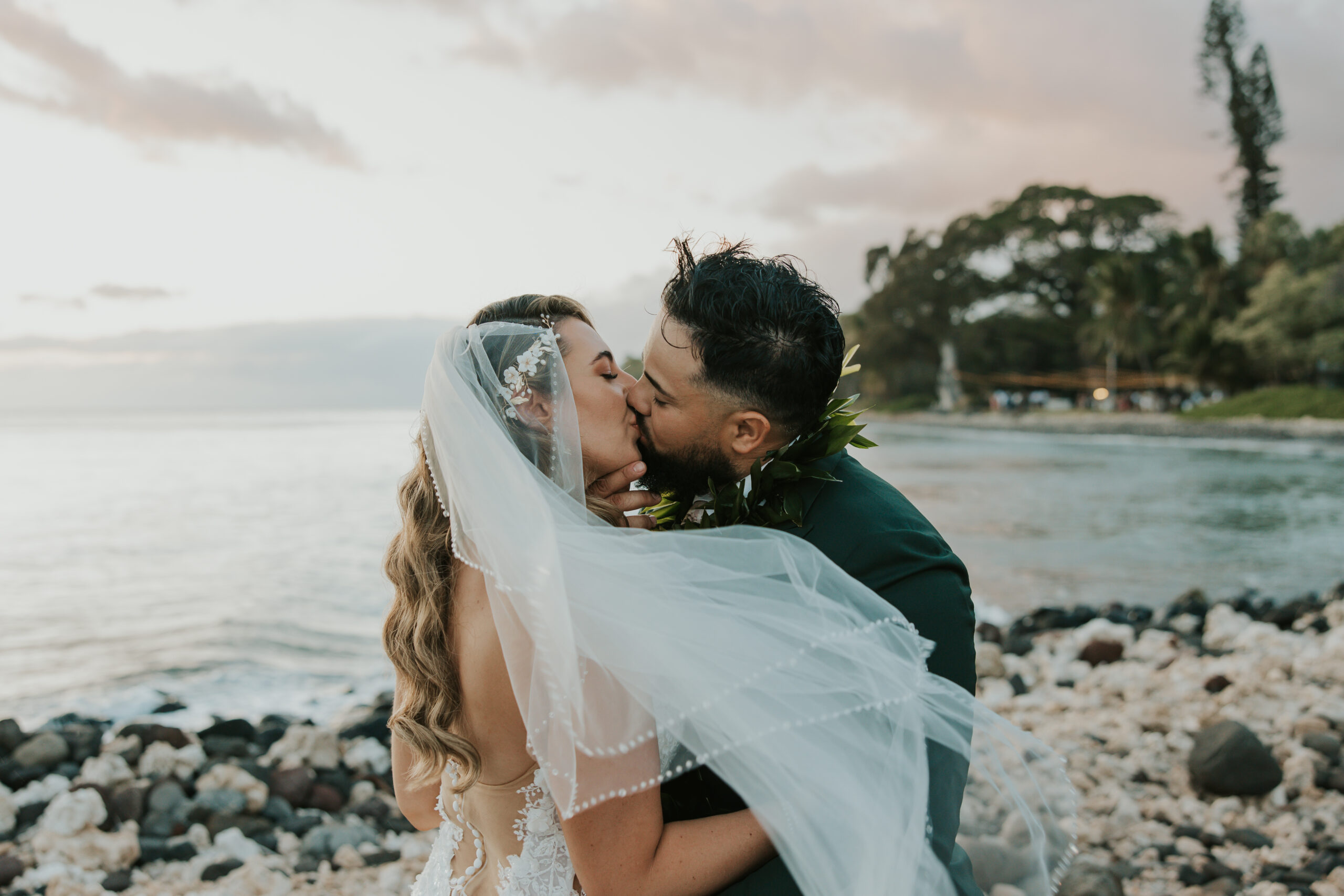 Bride and groom share a romantic kiss at sunset, her veil catching the breeze beside the rocky shore of a Maui wedding venue.