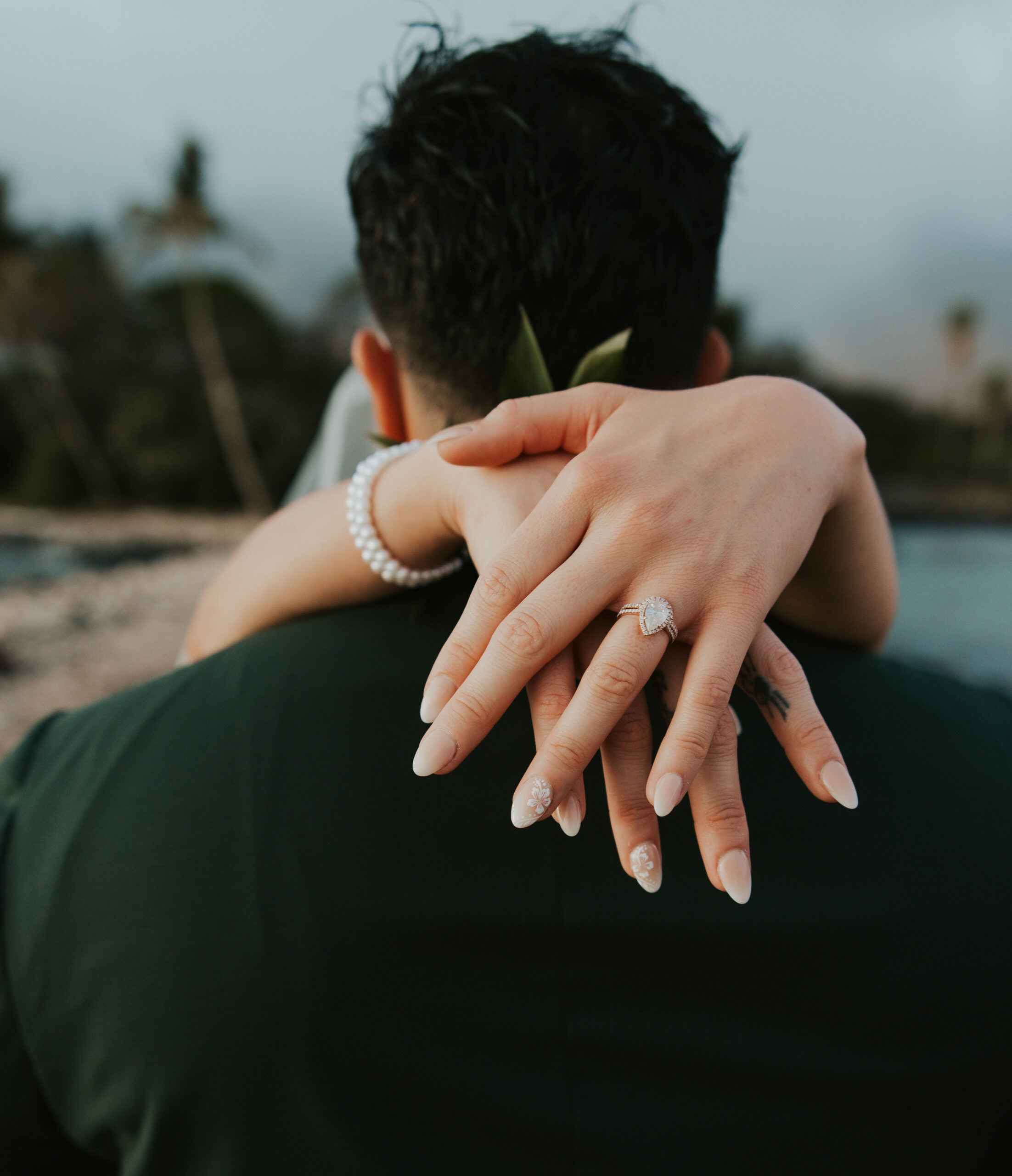 Close-up of the bride’s hands wrapped around the groom’s neck, showing off her sparkling heart-shaped engagement ring and pearl bracelet.
