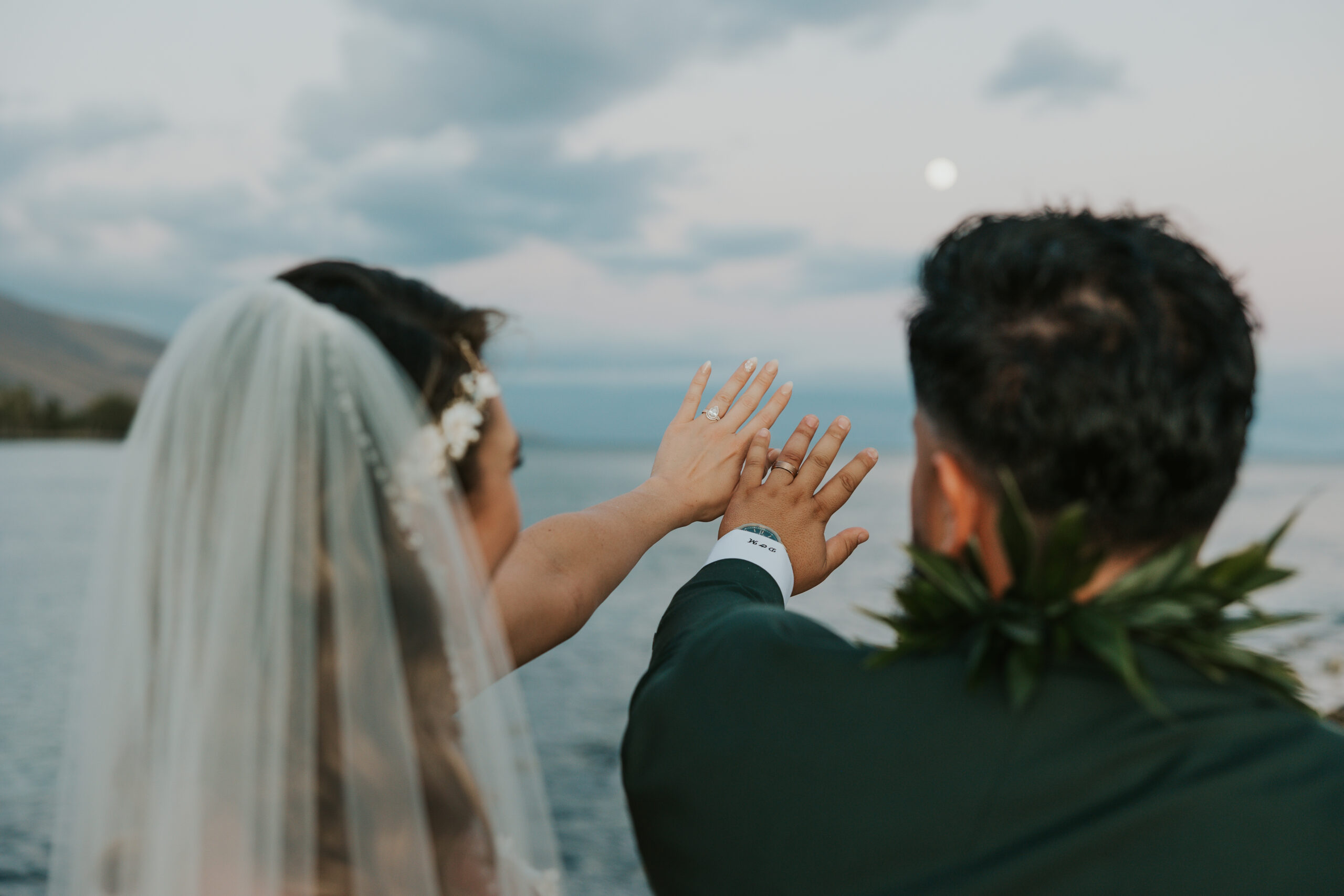 Couple raise their hands toward the moonlight, proudly showing off their wedding bands at a scenic Maui wedding venue.
