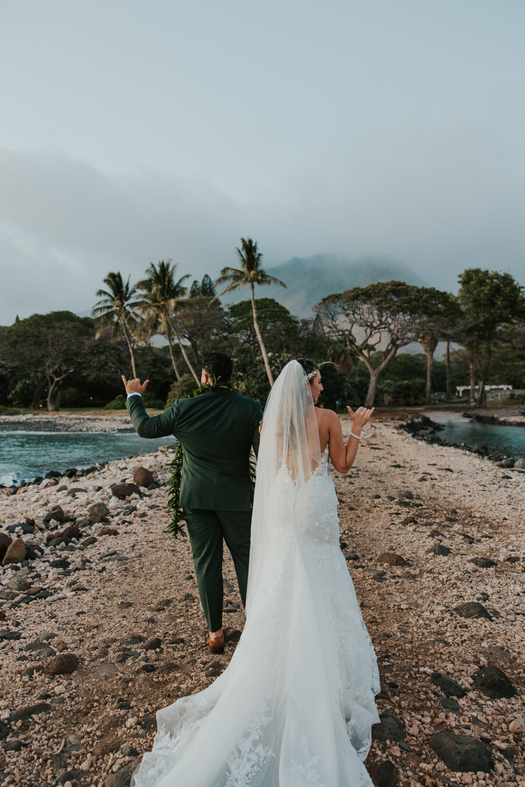 Bride and groom walking hand-in-hand down a rocky path by the ocean, throwing shaka signs in celebration at a Maui wedding venue.