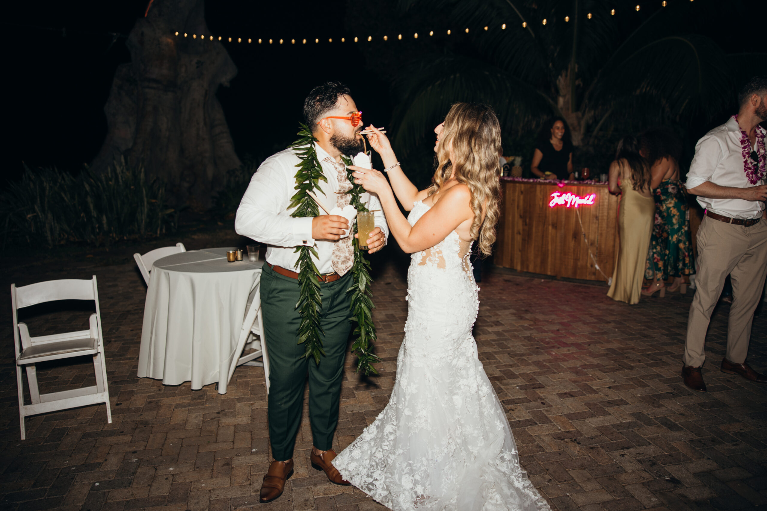 Bride feeds the groom during the nighttime celebration, surrounded by laughter, string lights, and a glowing “Just Mauied” sign.
