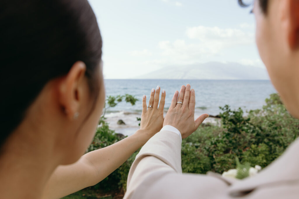 Newlyweds holding up their wedding rings with the ocean and neighboring island visible in the distance.