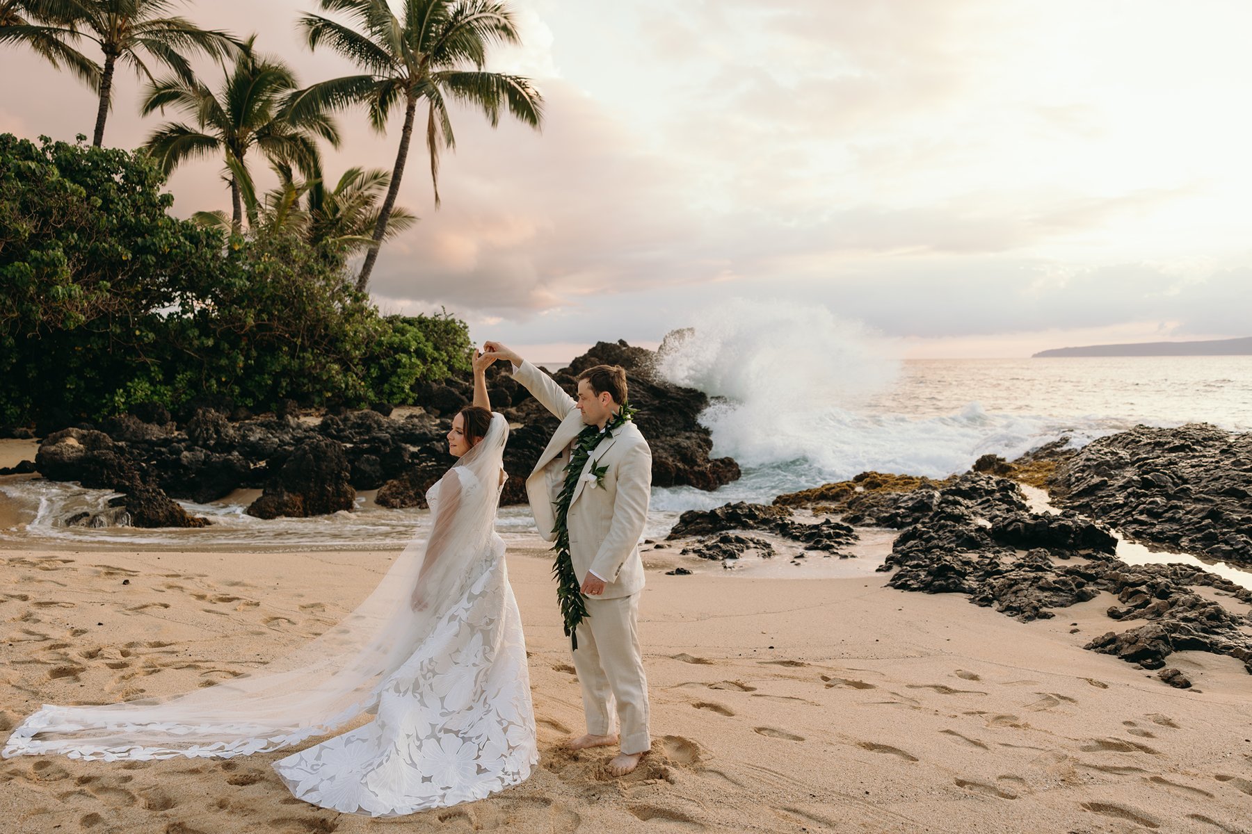 Groom twirling his bride on a sandy Maui beach with palm trees and ocean waves during sunset elopement photography.