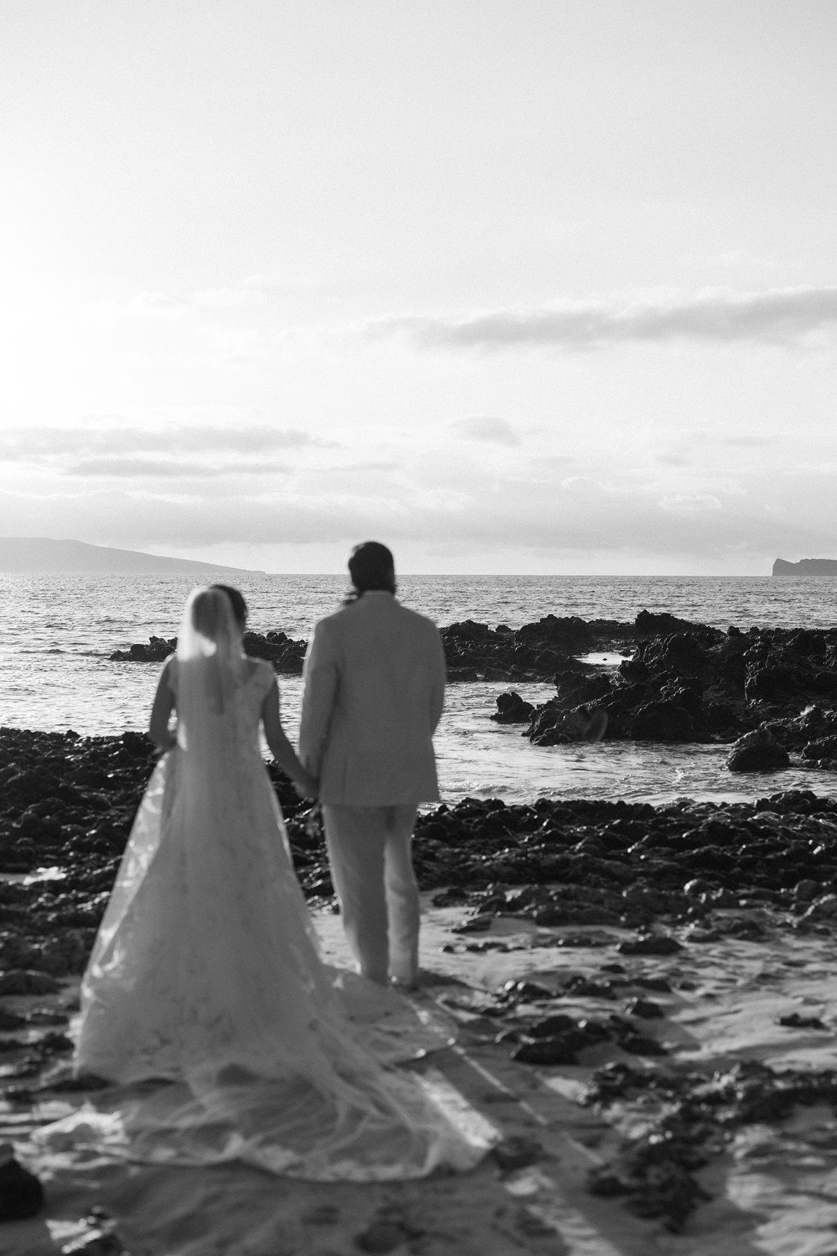 Black and white photo of newlyweds walking together along rocky Maui shoreline at sunset