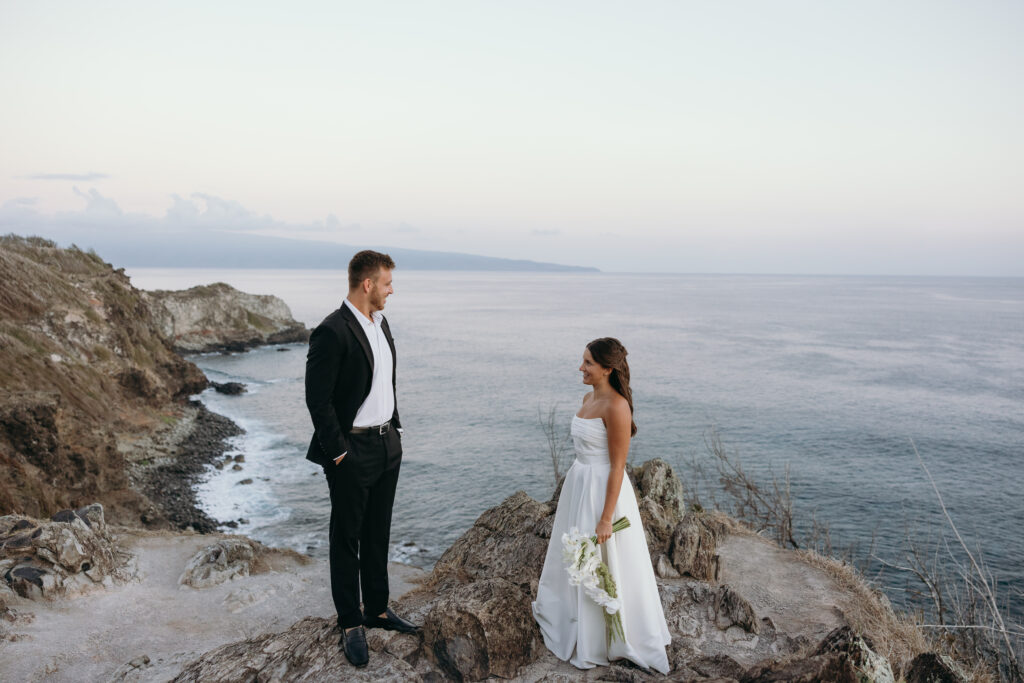 Bride and groom stand on rocky cliffs above the ocean during golden hour, showcased with drone videography.