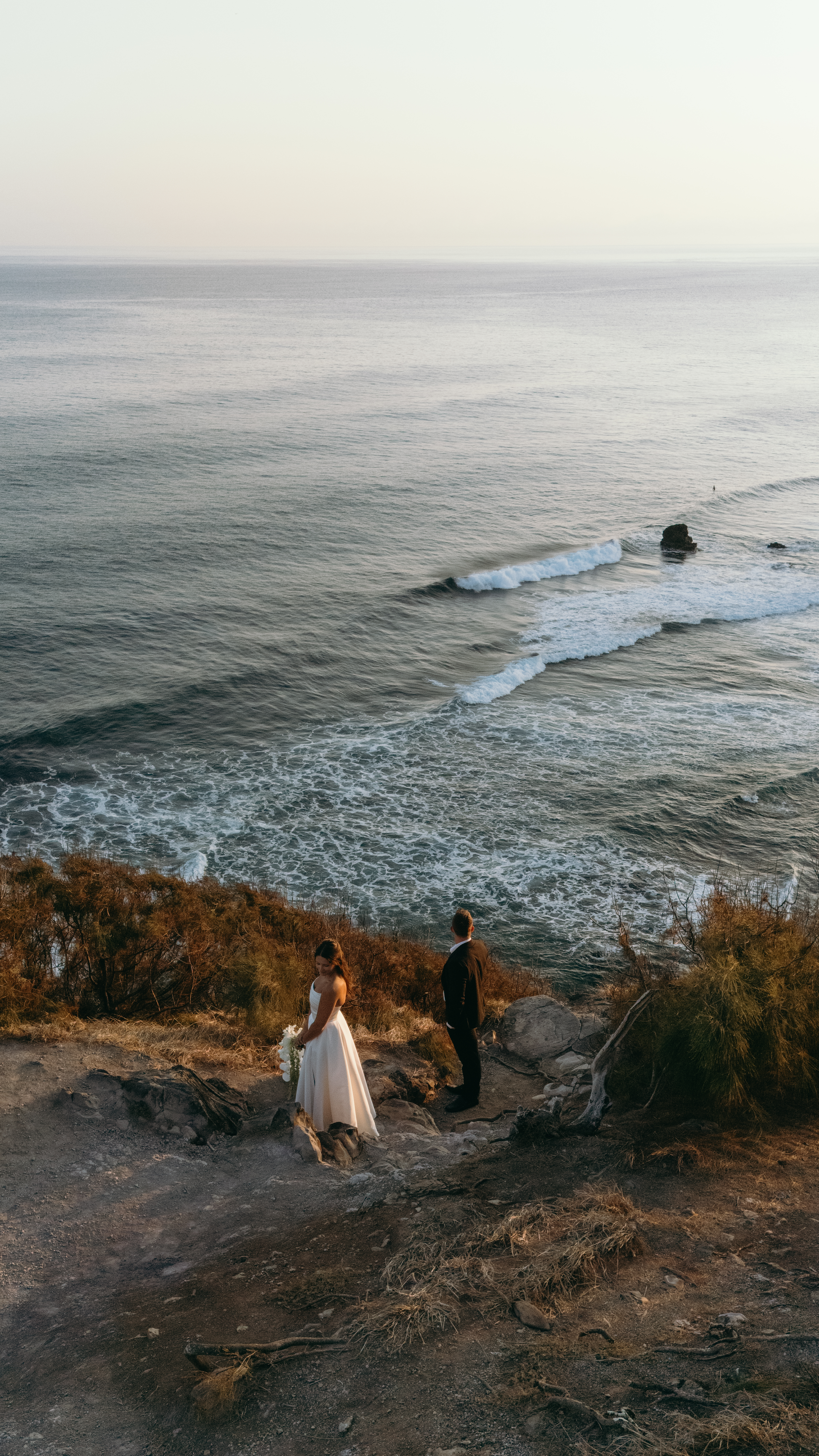 Bride and groom pause on a cliffside trail overlooking rolling waves and dramatic coastline, documented with drone videography.