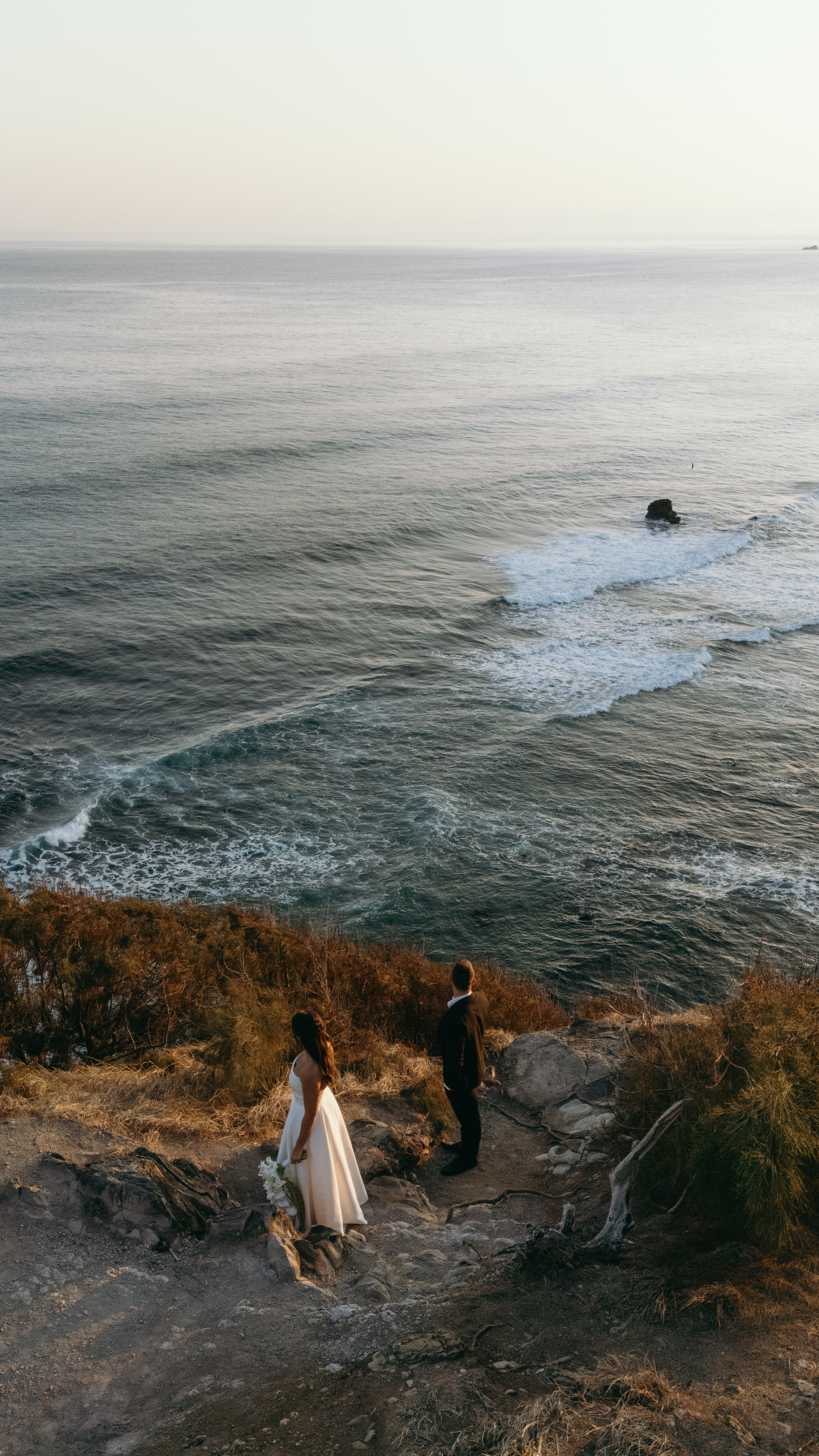 Bride and groom walk along a cliffside trail overlooking crashing waves in West Maui, captured with drone videography.
