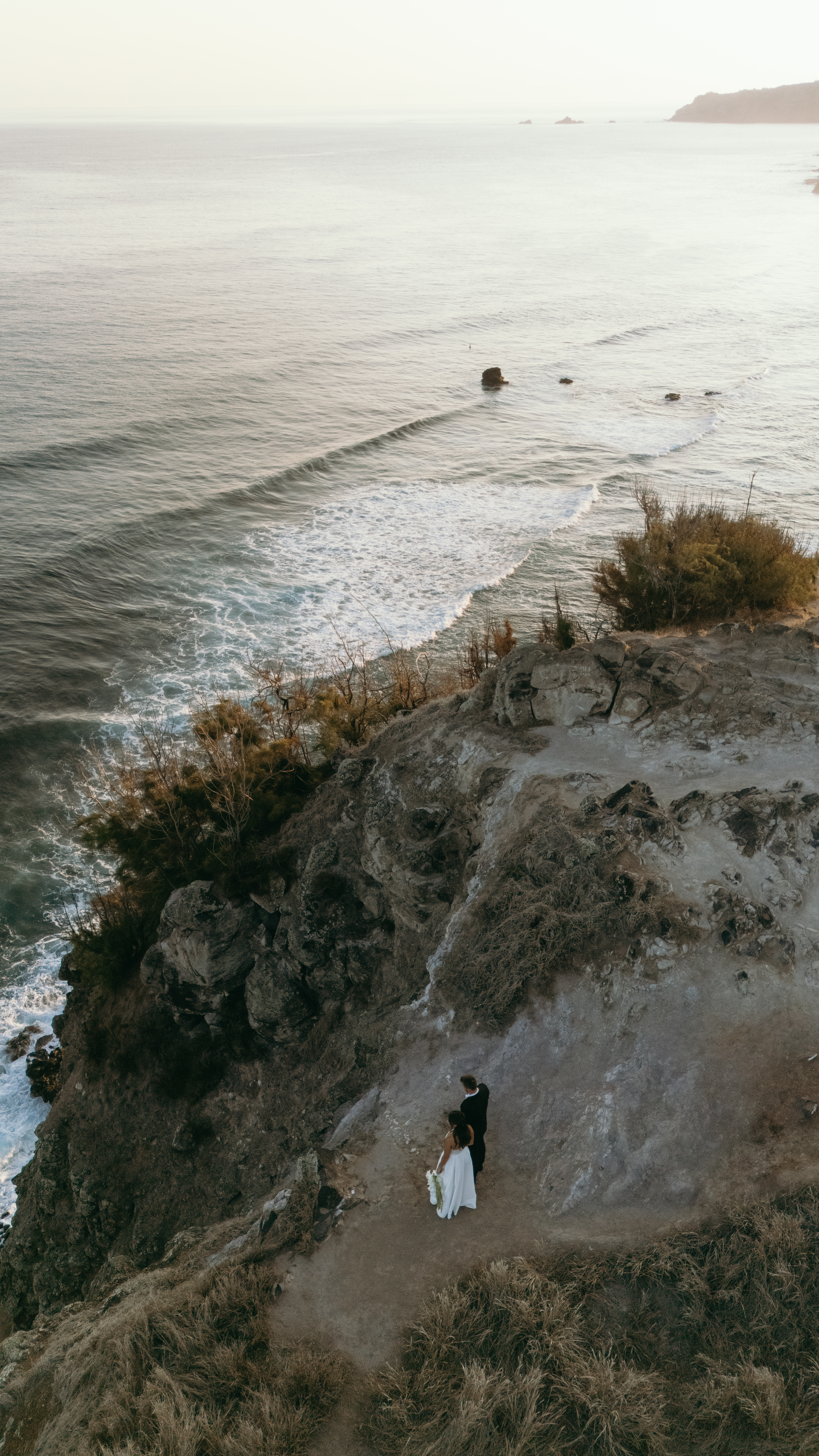 High-angle view of a bride and groom on a narrow coastal path above the ocean during a West Maui elopement.