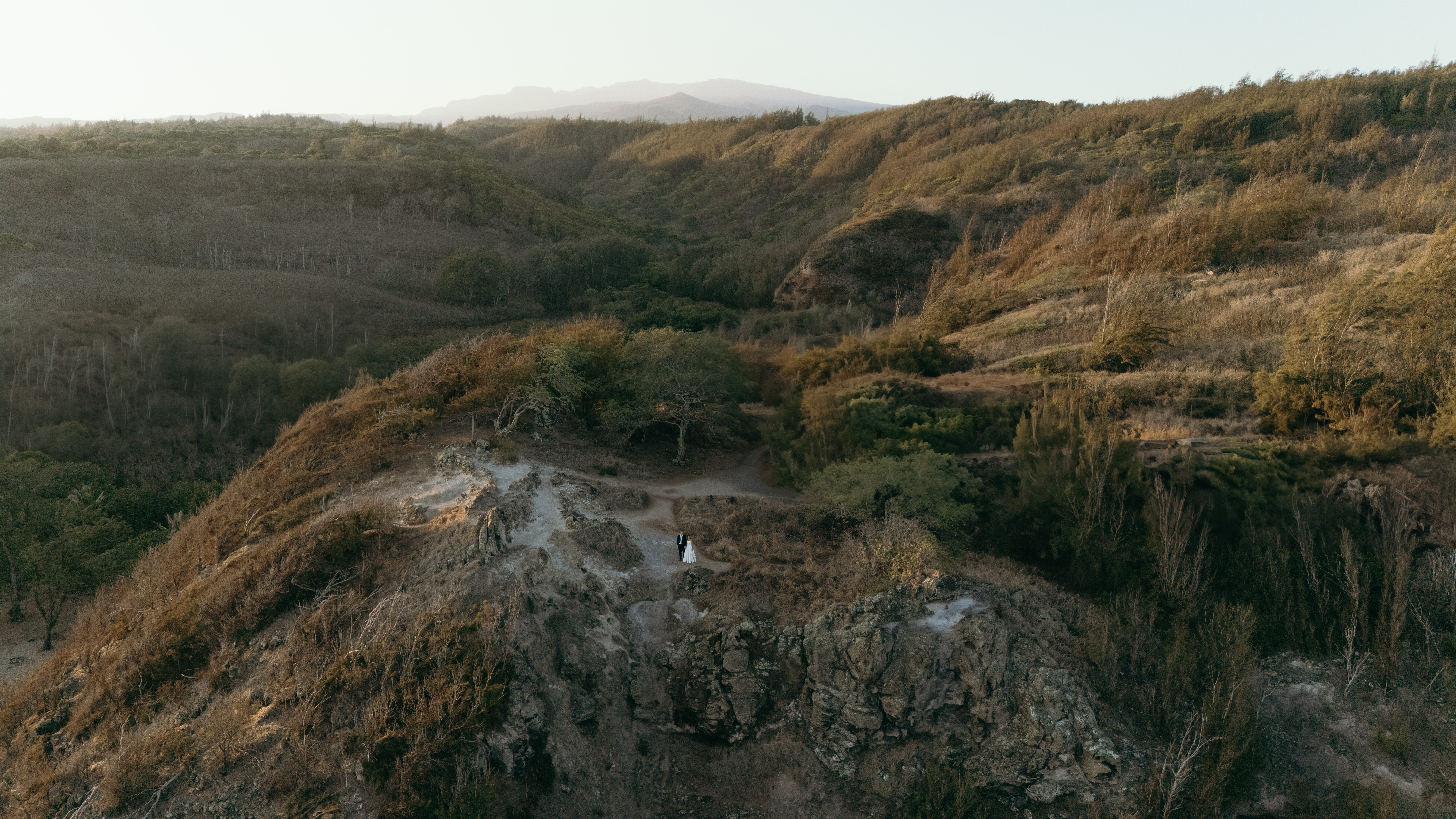 Aerial view of a couple standing together on a rugged West Maui cliff surrounded by lush landscape, captured with drone videography.