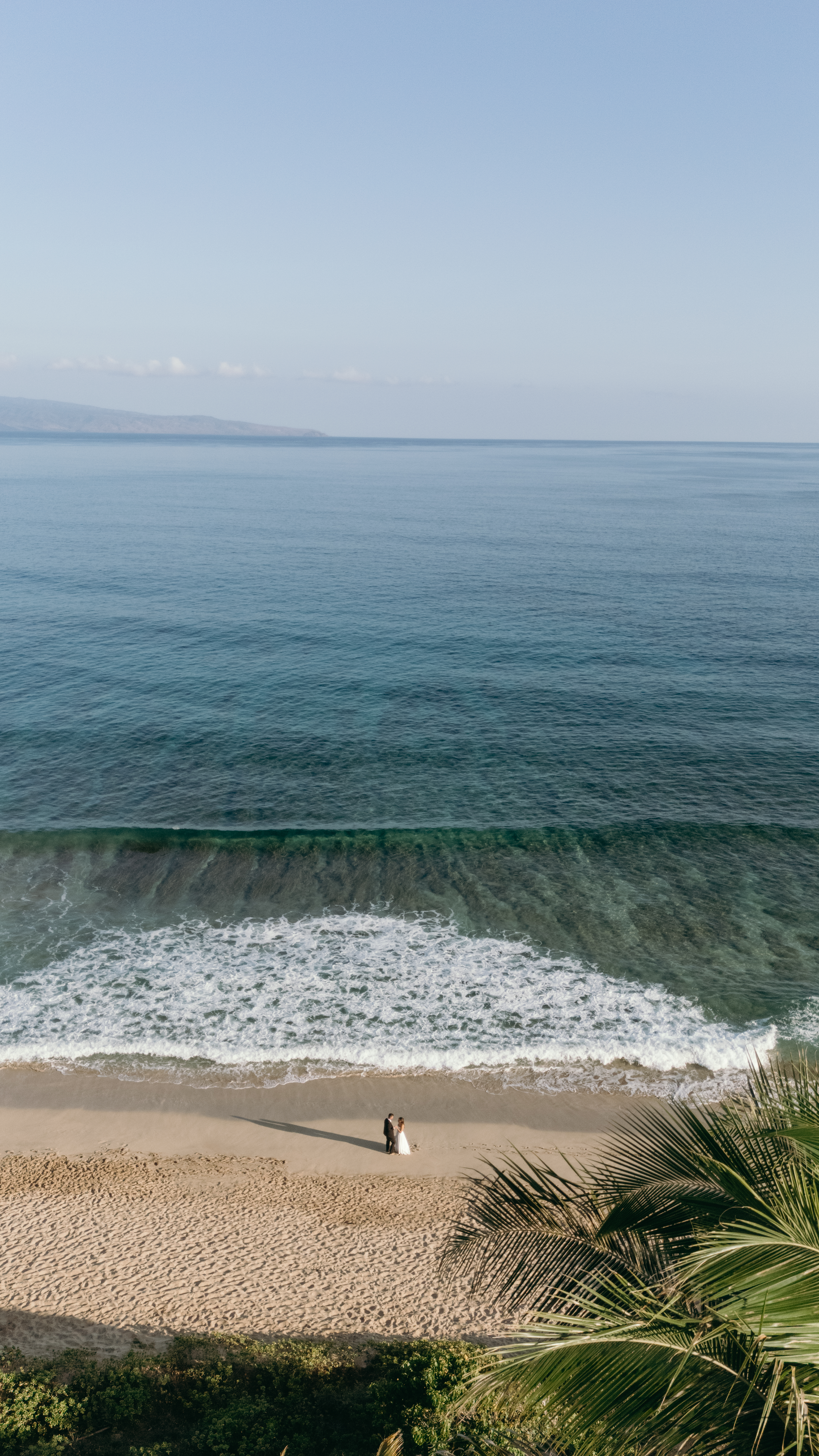 Aerial view of a couple standing on a secluded Maui beach with turquoise water and gentle waves, ideal for drone videography.