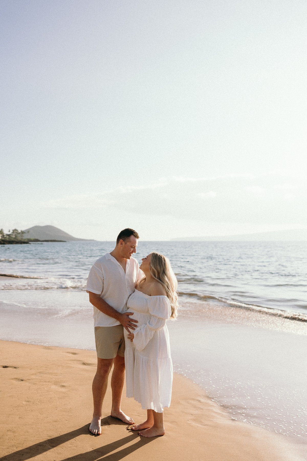 Couple standing together on a sandy beach holding baby bump during a Maui babymoon maternity photo session.