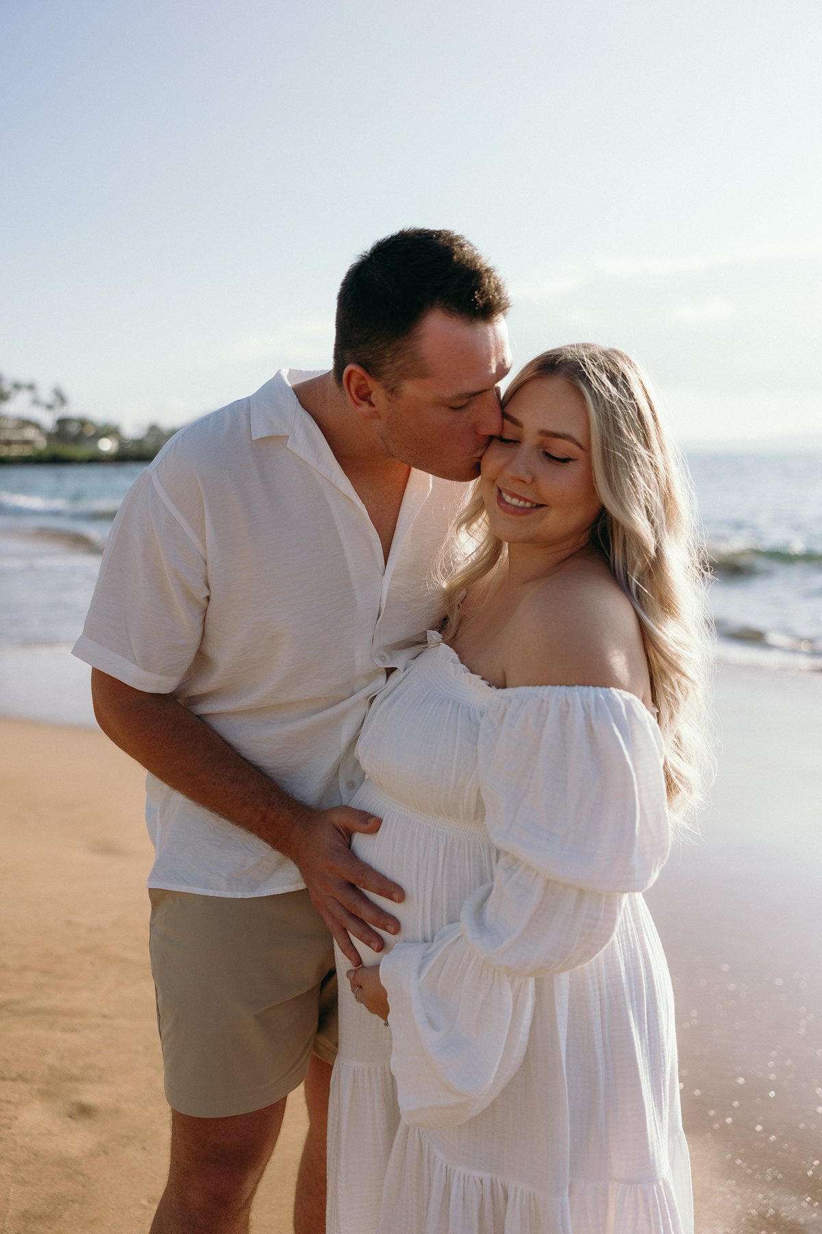 Couple embracing on the beach as the father kisses the mother’s forehead during their Maui babymoon.