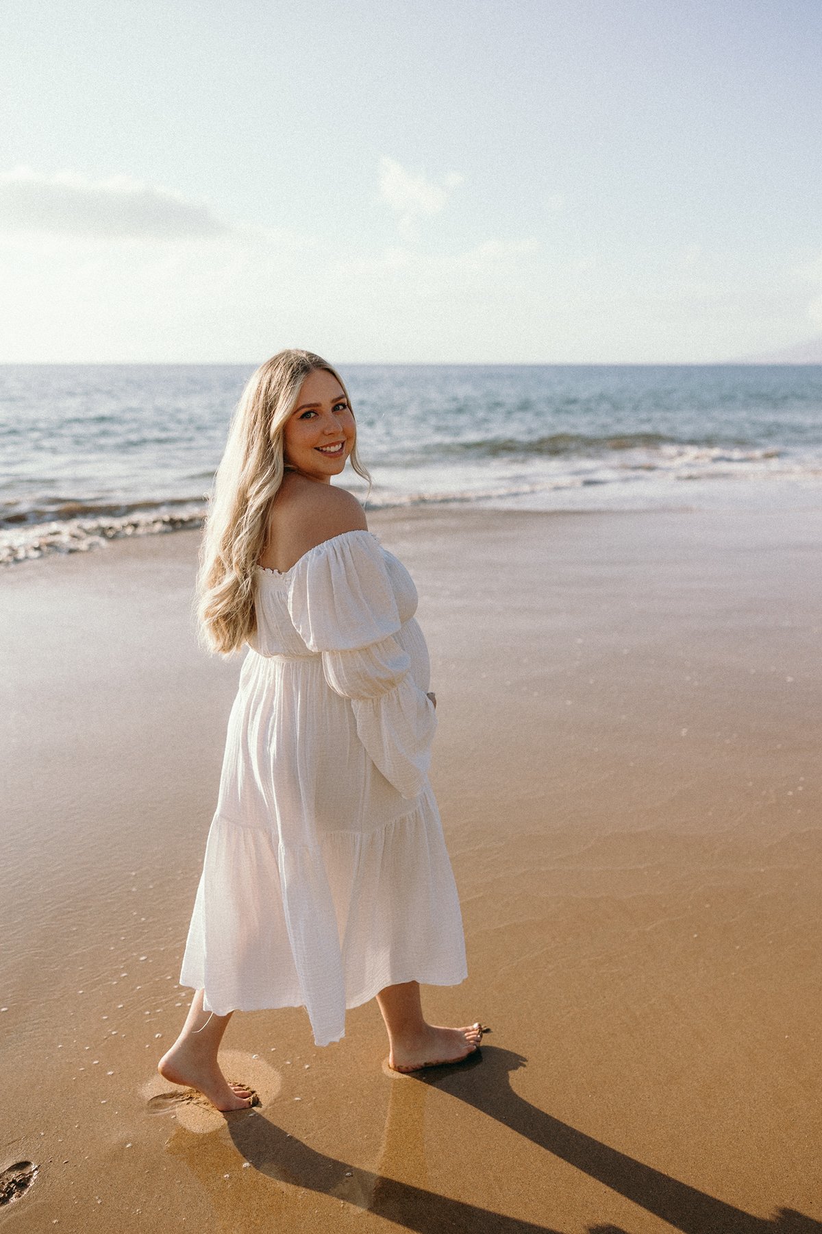 Pregnant woman walking barefoot along a Maui beach in a white dress during a maternity photoshoot.