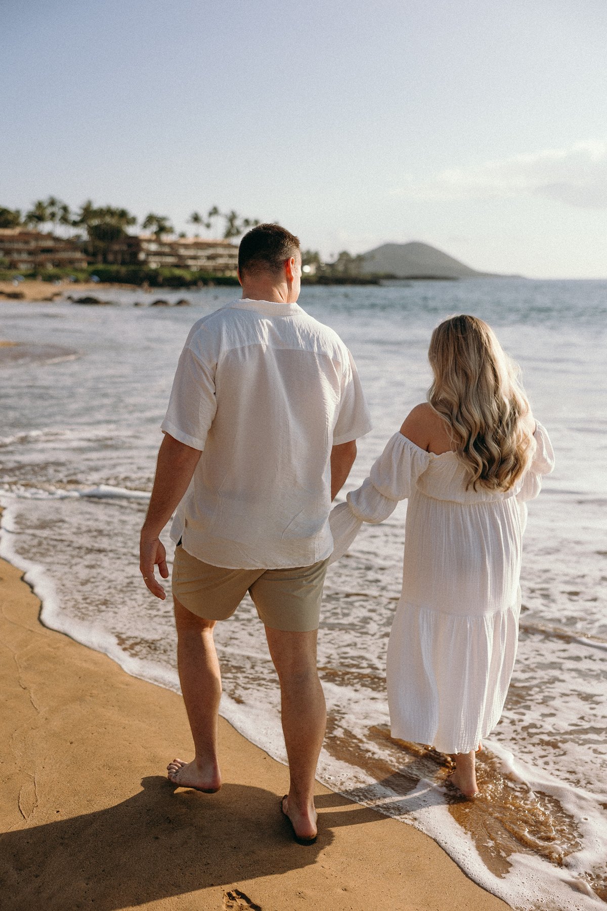 Expecting couple walking hand in hand along the shoreline during a peaceful Maui babymoon.