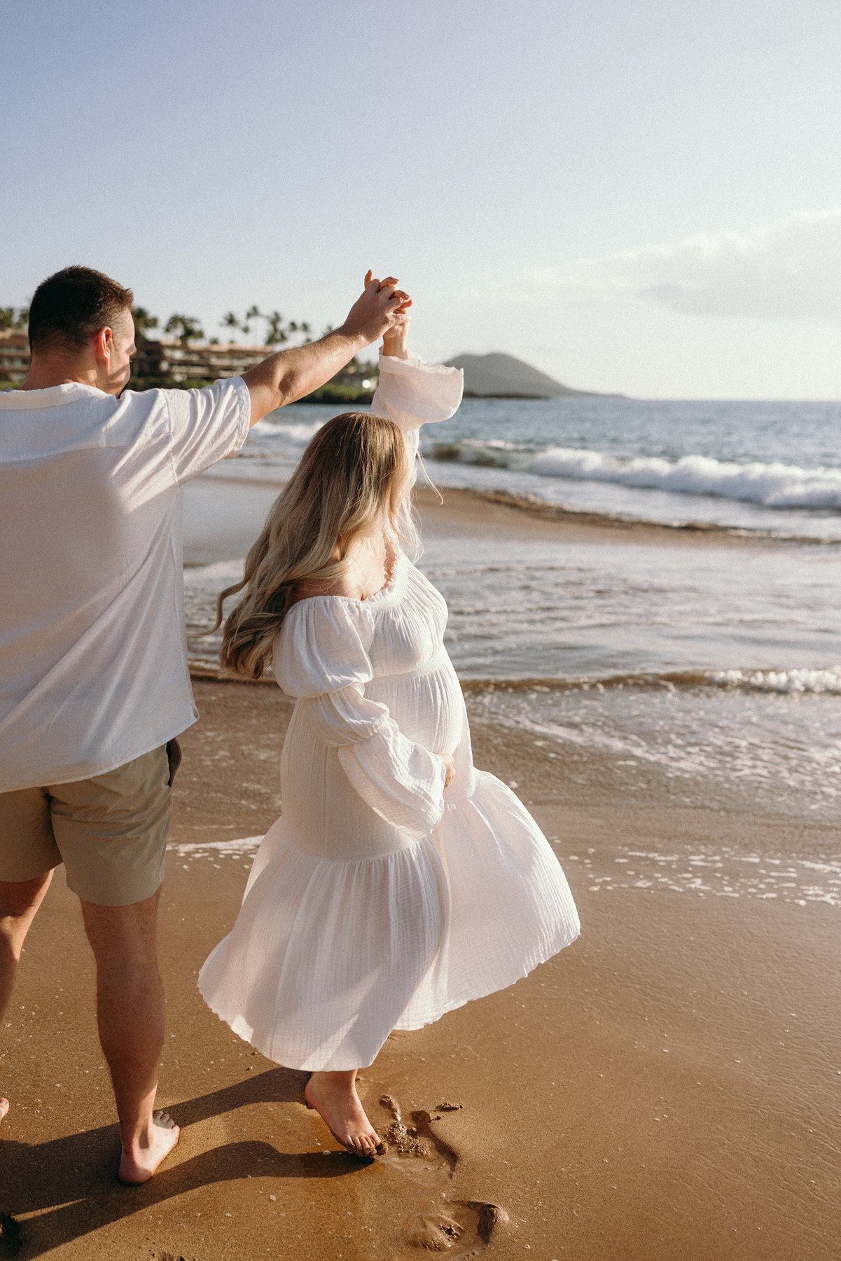 Expecting couple twirling together while walking along the beach during their Maui babymoon.