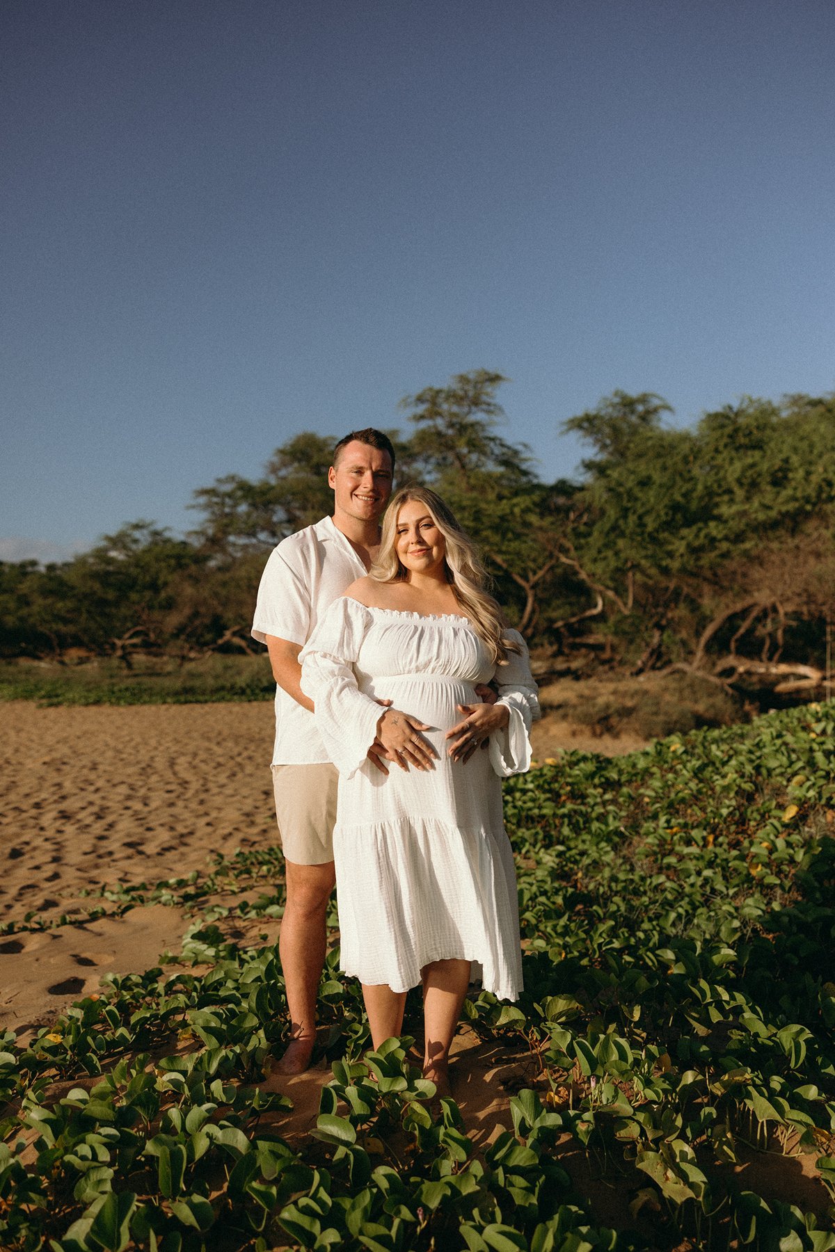 Pregnant woman and partner standing together on a sandy Maui beach surrounded by coastal plants.