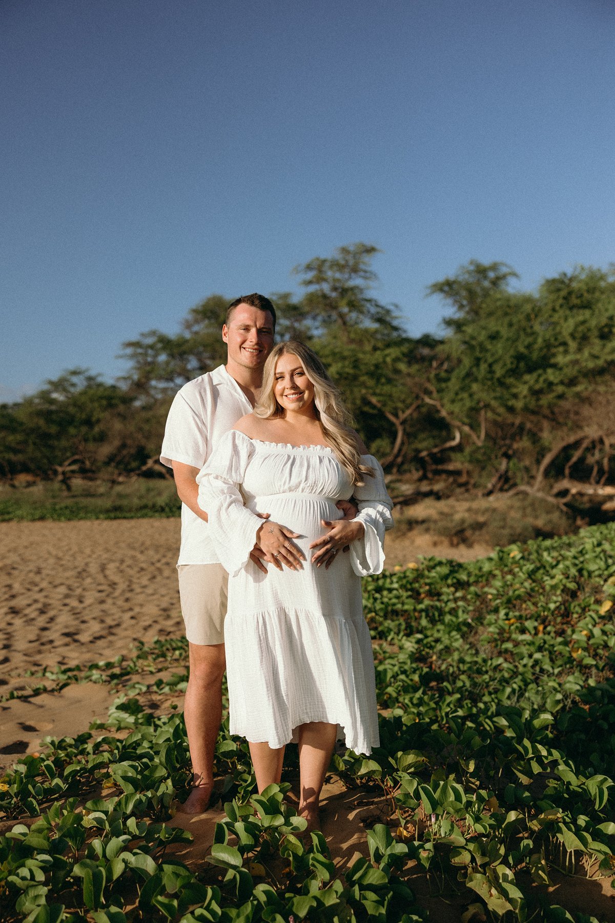 Expecting couple standing together among beach greenery during a relaxed maternity photo session in Maui.
