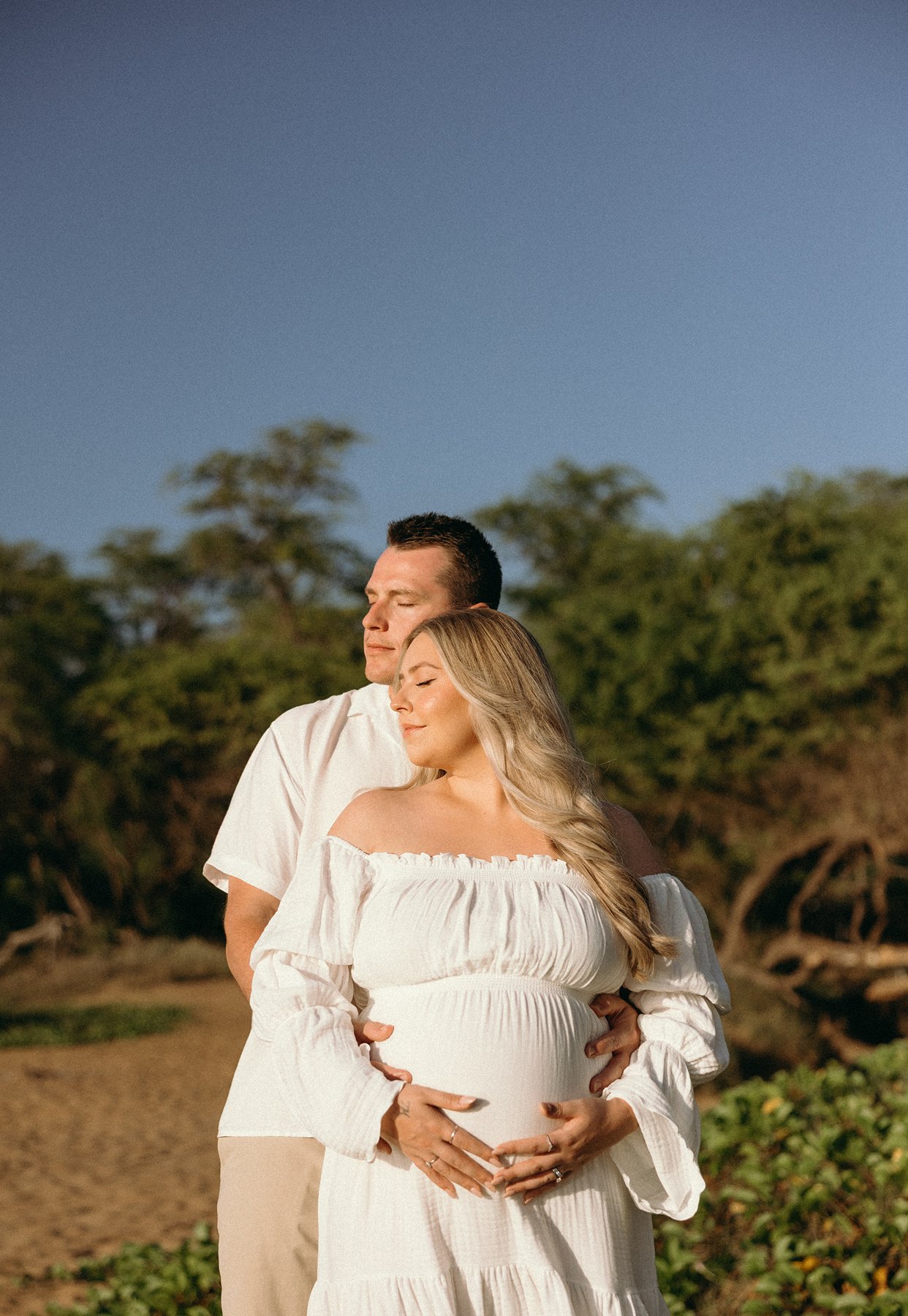 Expecting couple embracing with hands resting on baby bump during a beach maternity session in Maui.