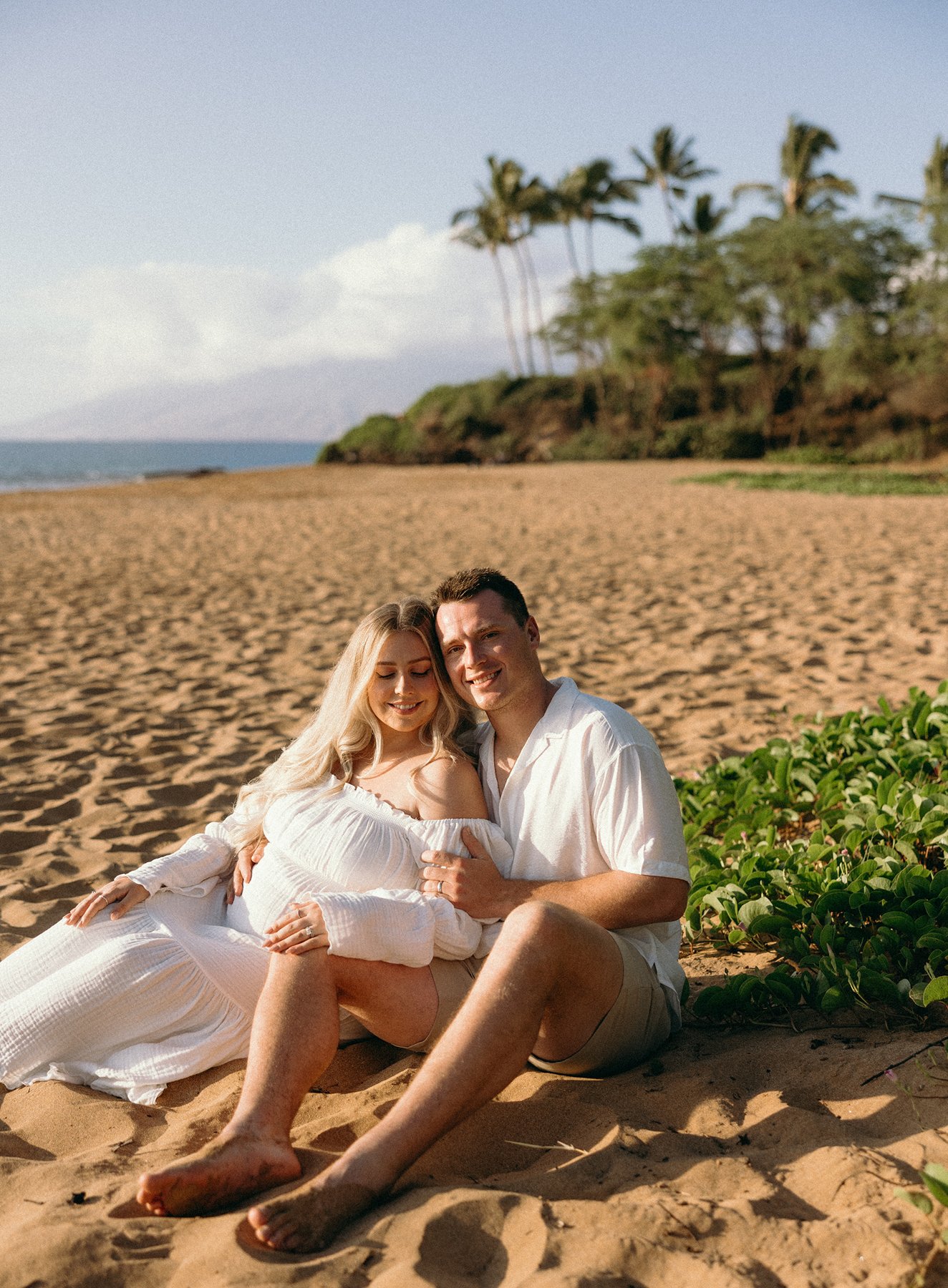 Expecting couple sitting together on a sandy Maui beach surrounded by tropical greenery during their Maui babymoon.