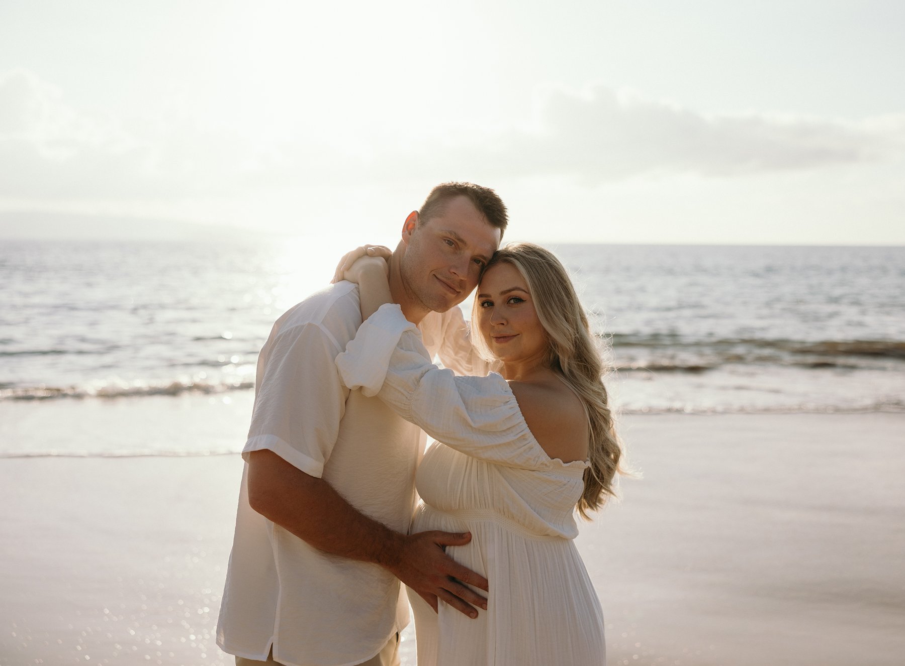 Expecting couple embracing on the beach with ocean waves behind them during golden hour.