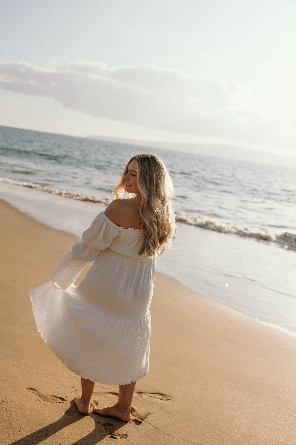 Pregnant woman walking barefoot along the shoreline with her dress flowing in the ocean breeze.