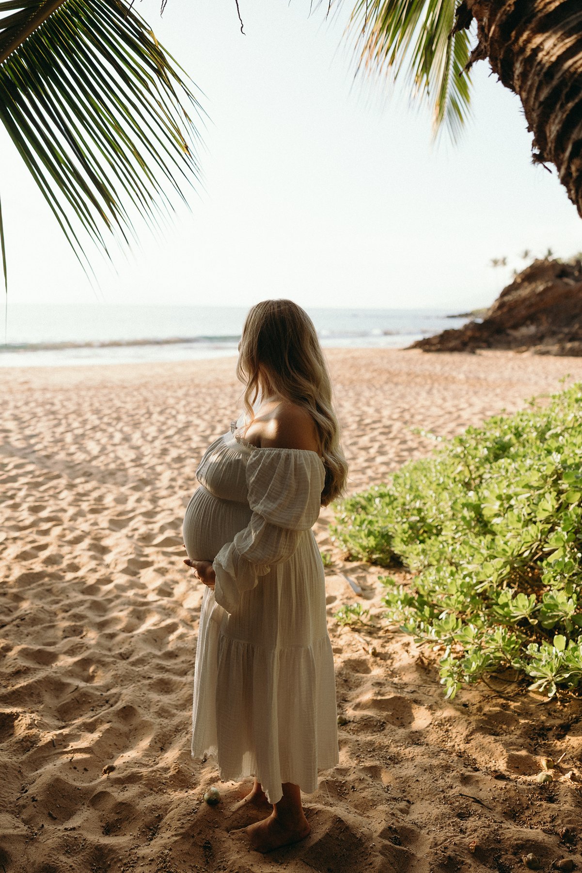 Pregnant woman standing beneath palm trees on a sandy beach during a peaceful Maui babymoon maternity session.