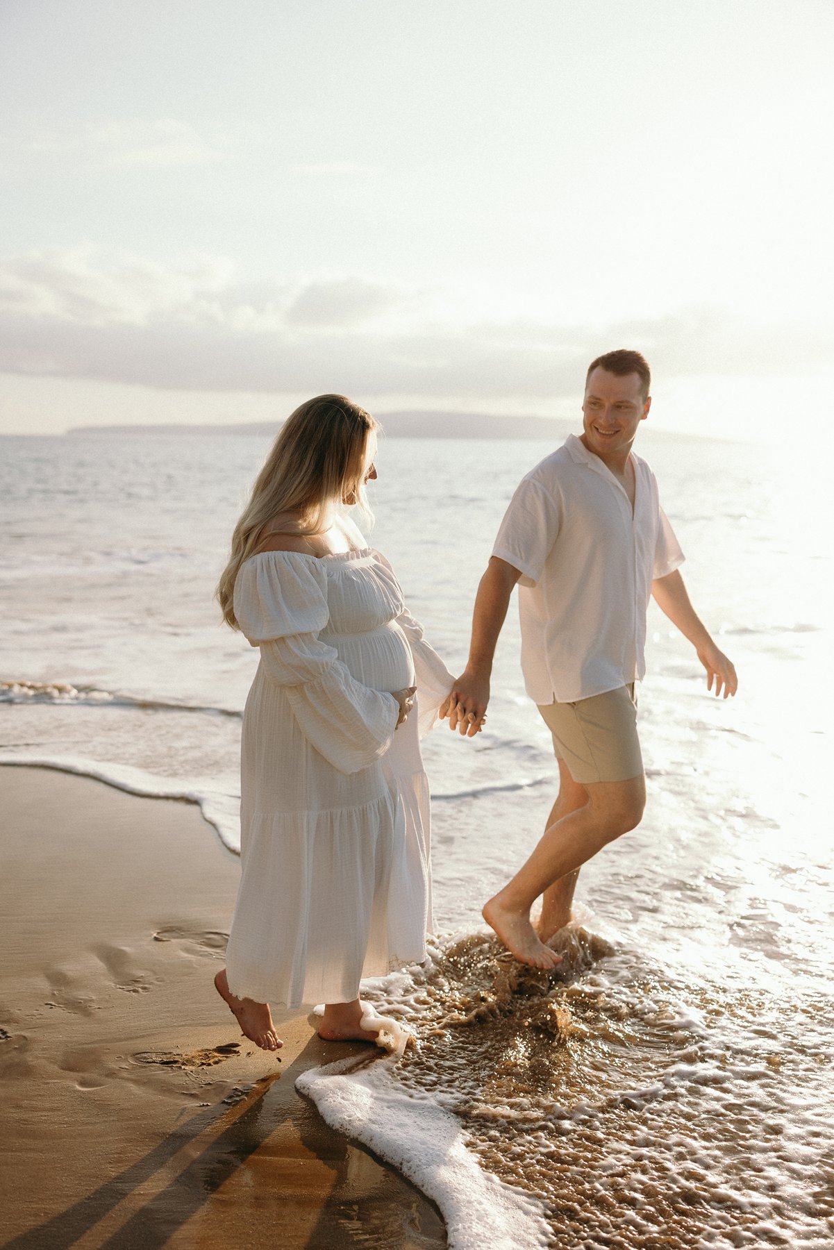 Expecting couple walking hand in hand through shallow ocean waves during golden hour maternity photos.