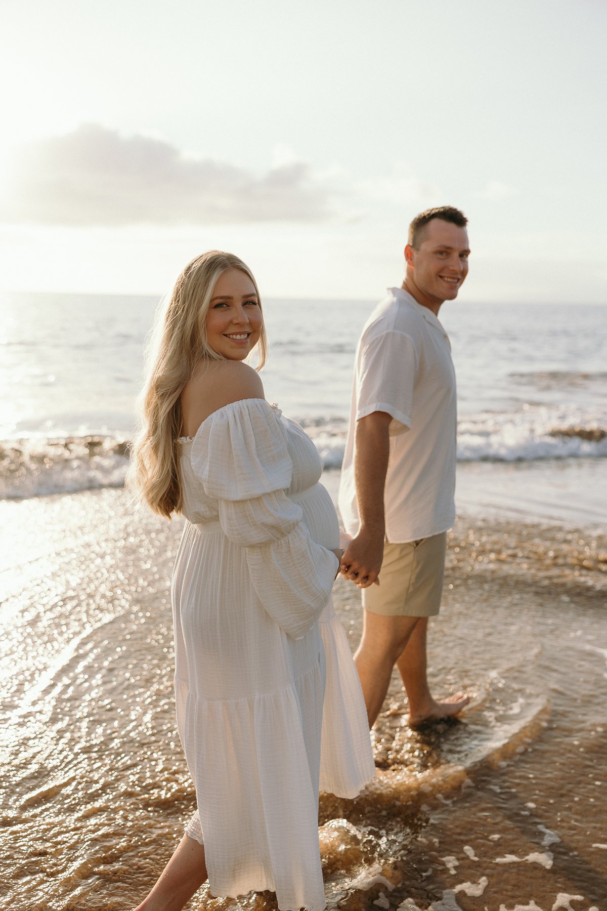 Pregnant woman smiling back while walking hand in hand with her partner through shallow ocean water.
