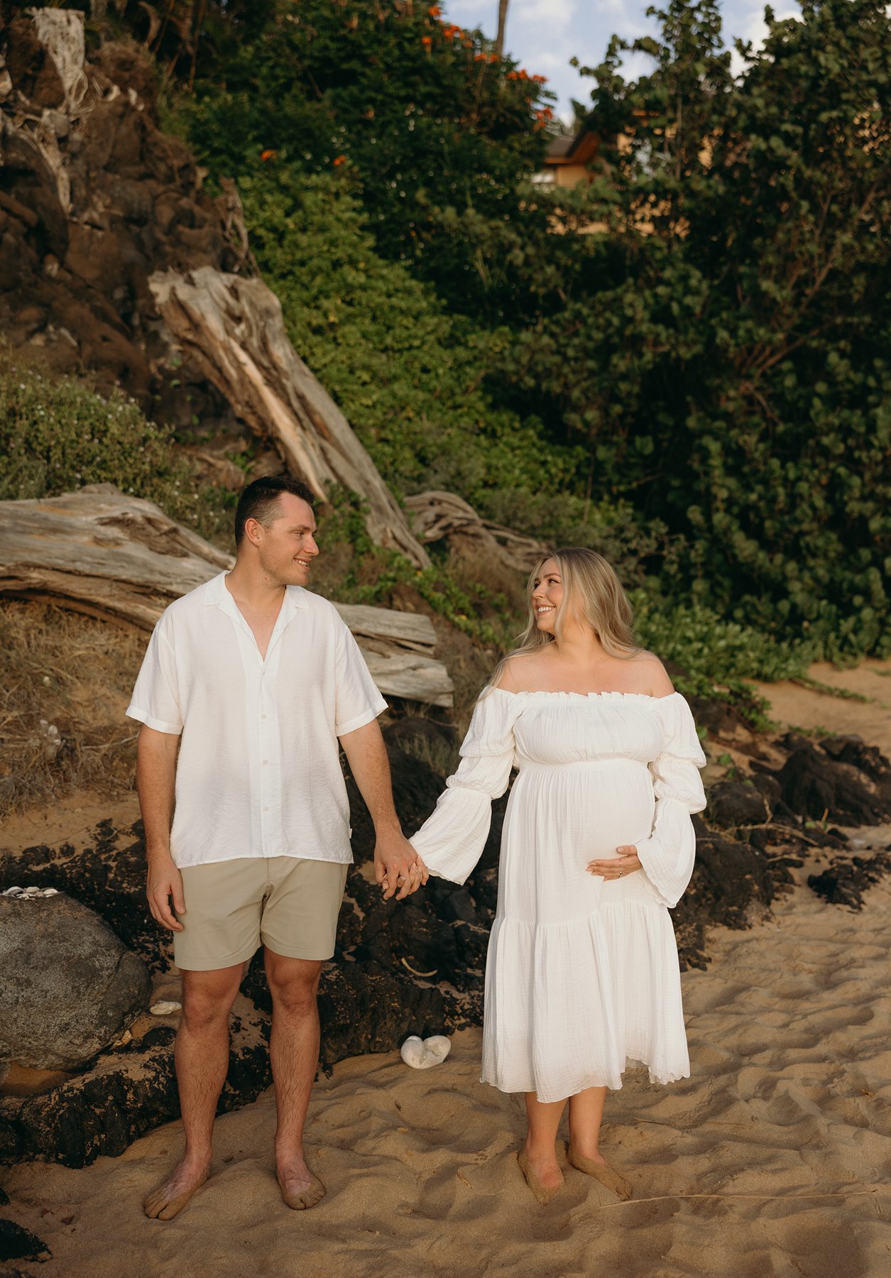 Pregnant couple holding hands on a sandy beach with lava rocks and tropical greenery in Maui.
