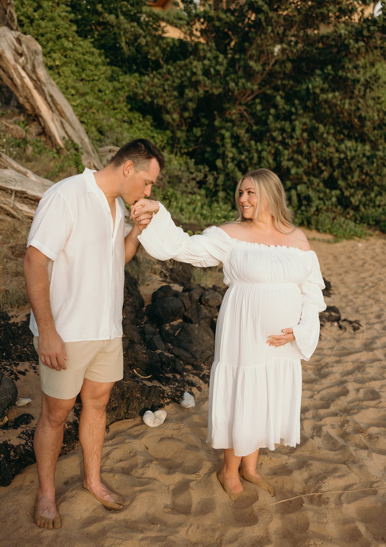 Father kissing the mother’s hand on a sandy Maui beach during their Maui babymoon maternity session.