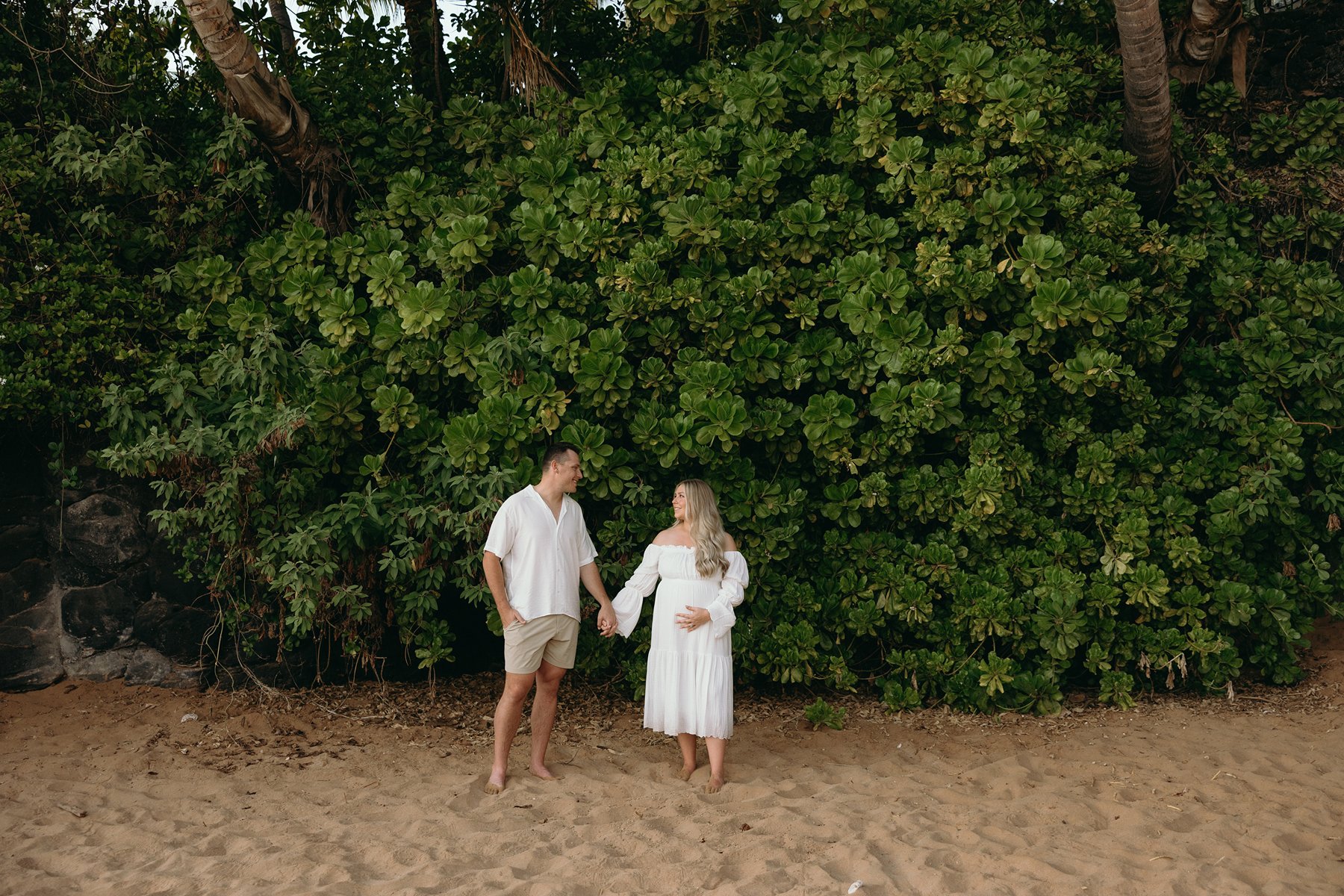 Pregnant couple holding hands in front of lush beach plants during a relaxed Maui babymoon photoshoot.