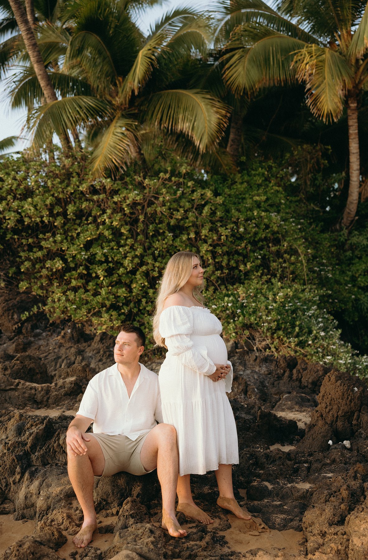 Expecting couple sitting on lava rocks beneath palm trees during a Maui babymoon maternity photo session.