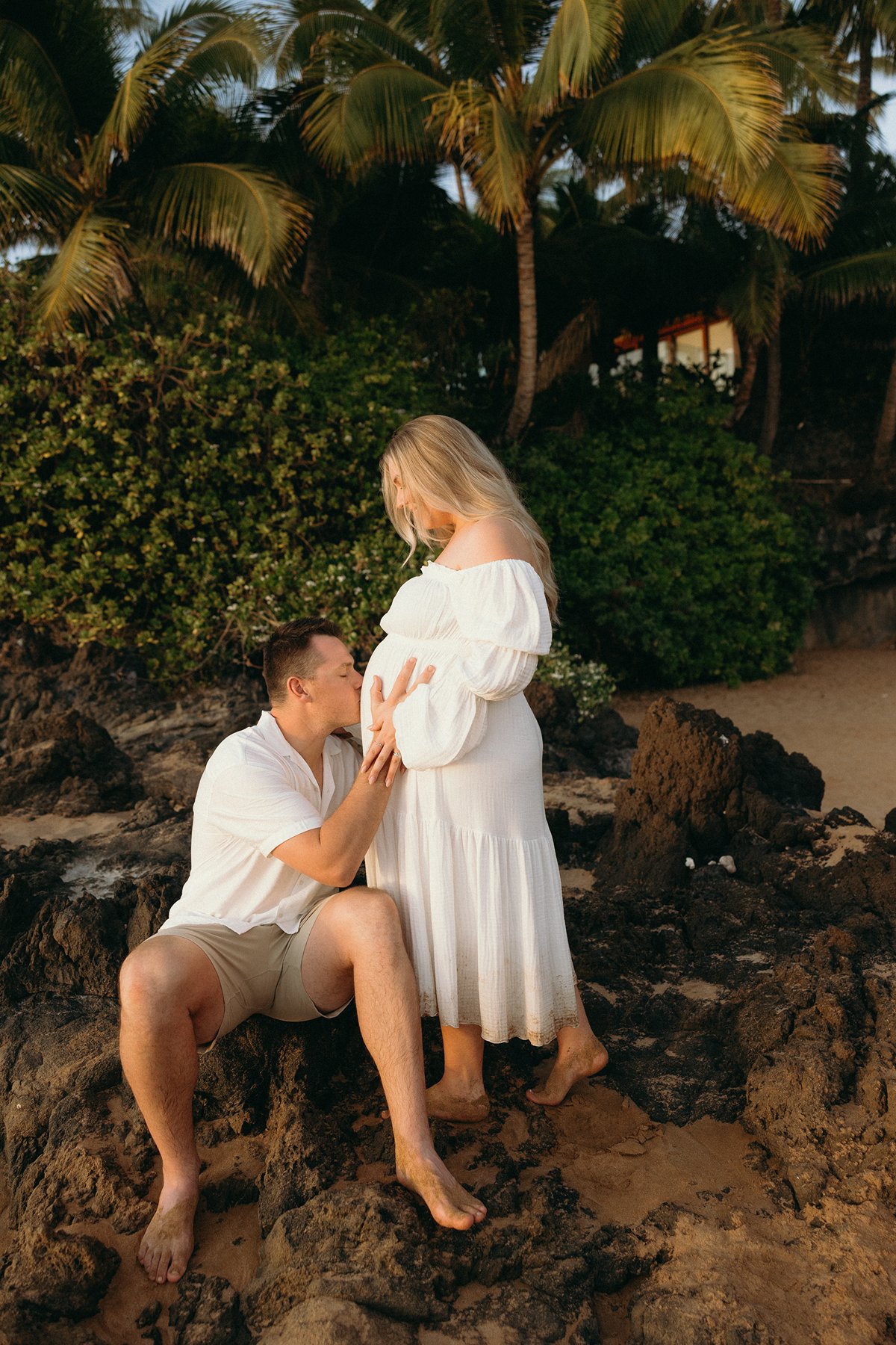 Expecting father kissing baby bump while sitting on lava rocks during a Maui babymoon maternity photo session.