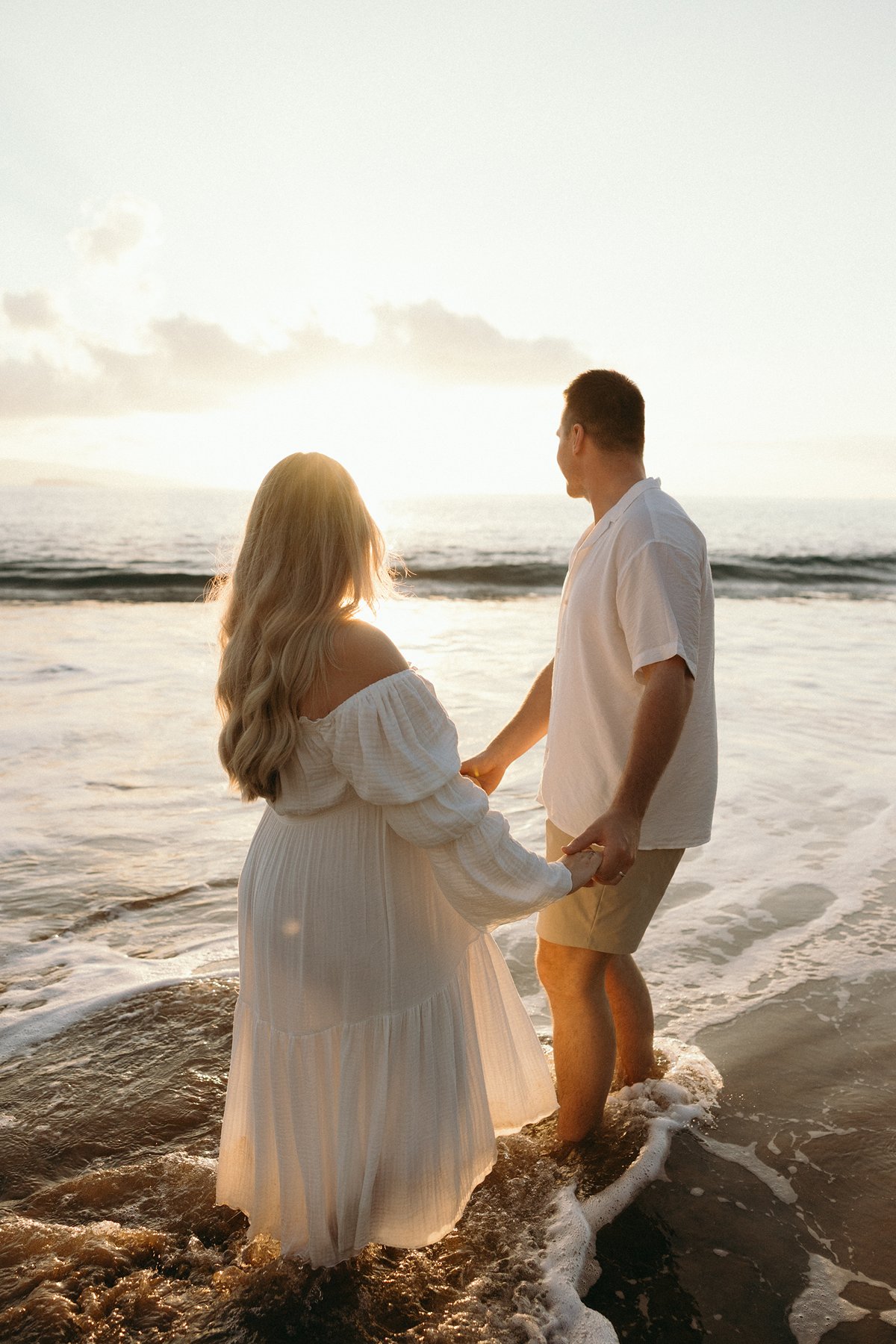 Couple holding hands while standing in gentle ocean waves during their Maui babymoon maternity session.