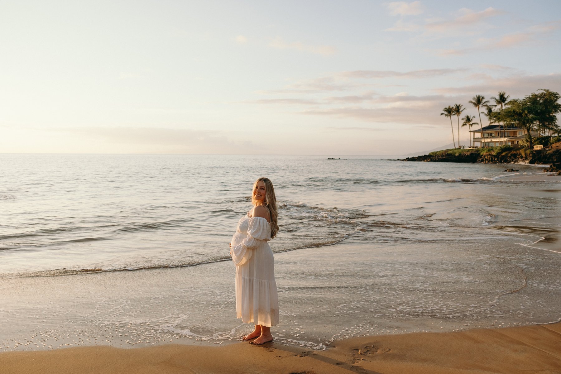 Pregnant woman standing along the shoreline at sunset with gentle waves rolling in.