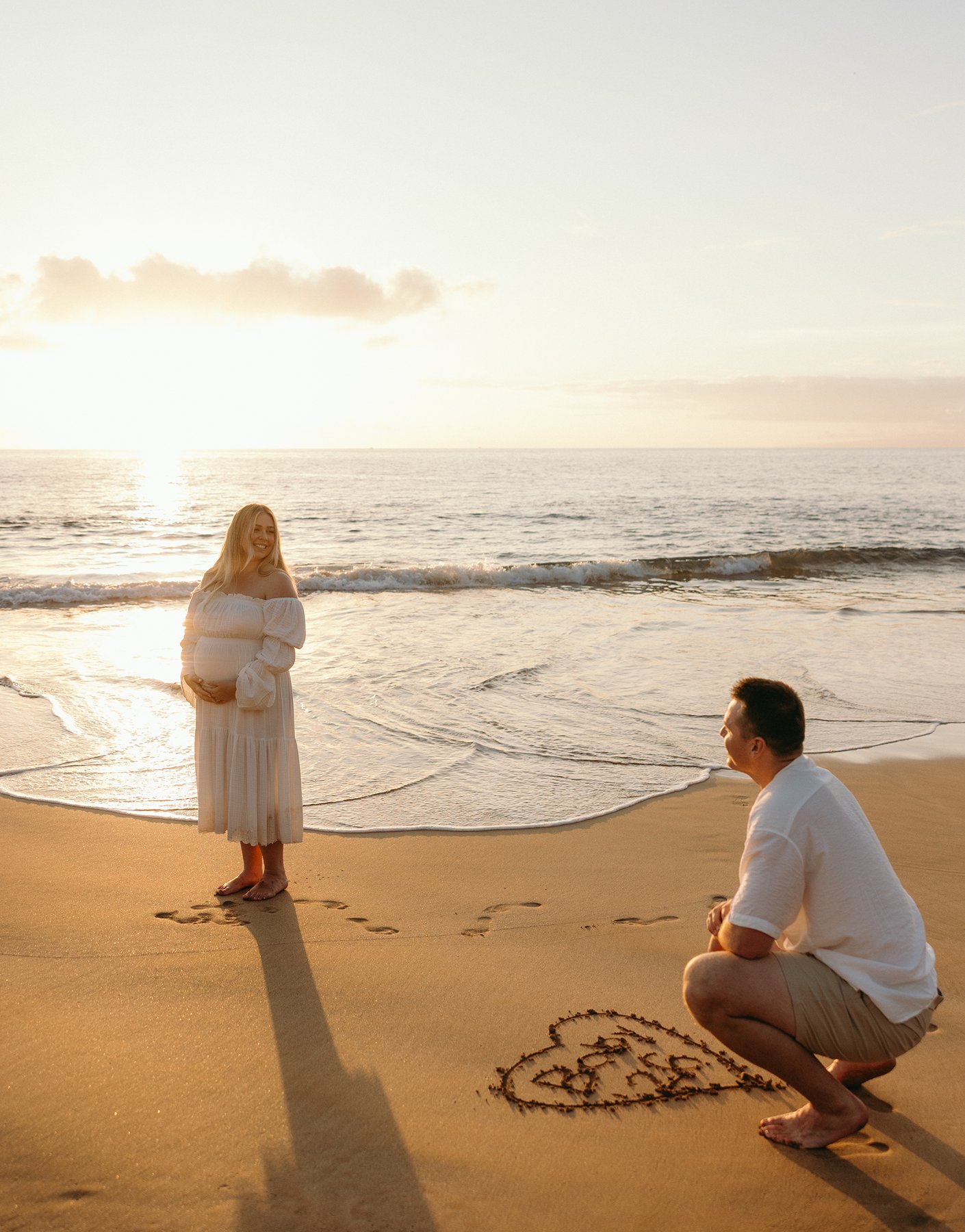 Expecting mother smiling at her partner as he draws a heart in the sand during their Maui babymoon.