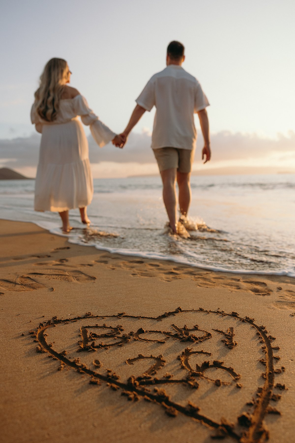 Couple walking along the beach at sunset with a heart drawn in the sand during their Maui babymoon.