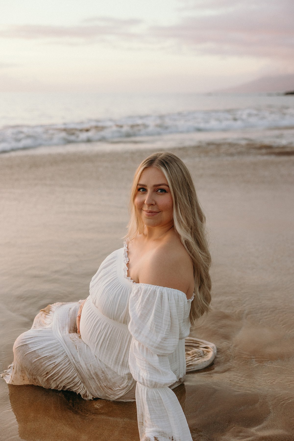Pregnant woman sitting on the shoreline in a white dress during a beach maternity photoshoot in Maui.