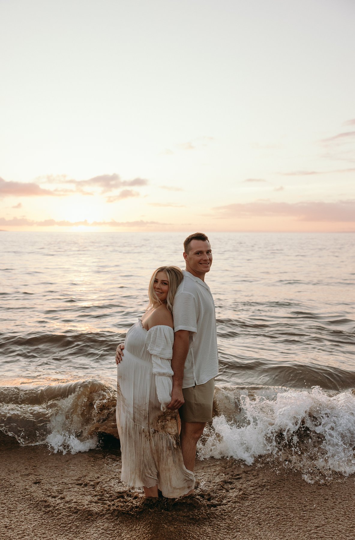 Expecting couple standing in gentle waves at sunset during their Maui babymoon maternity photos.