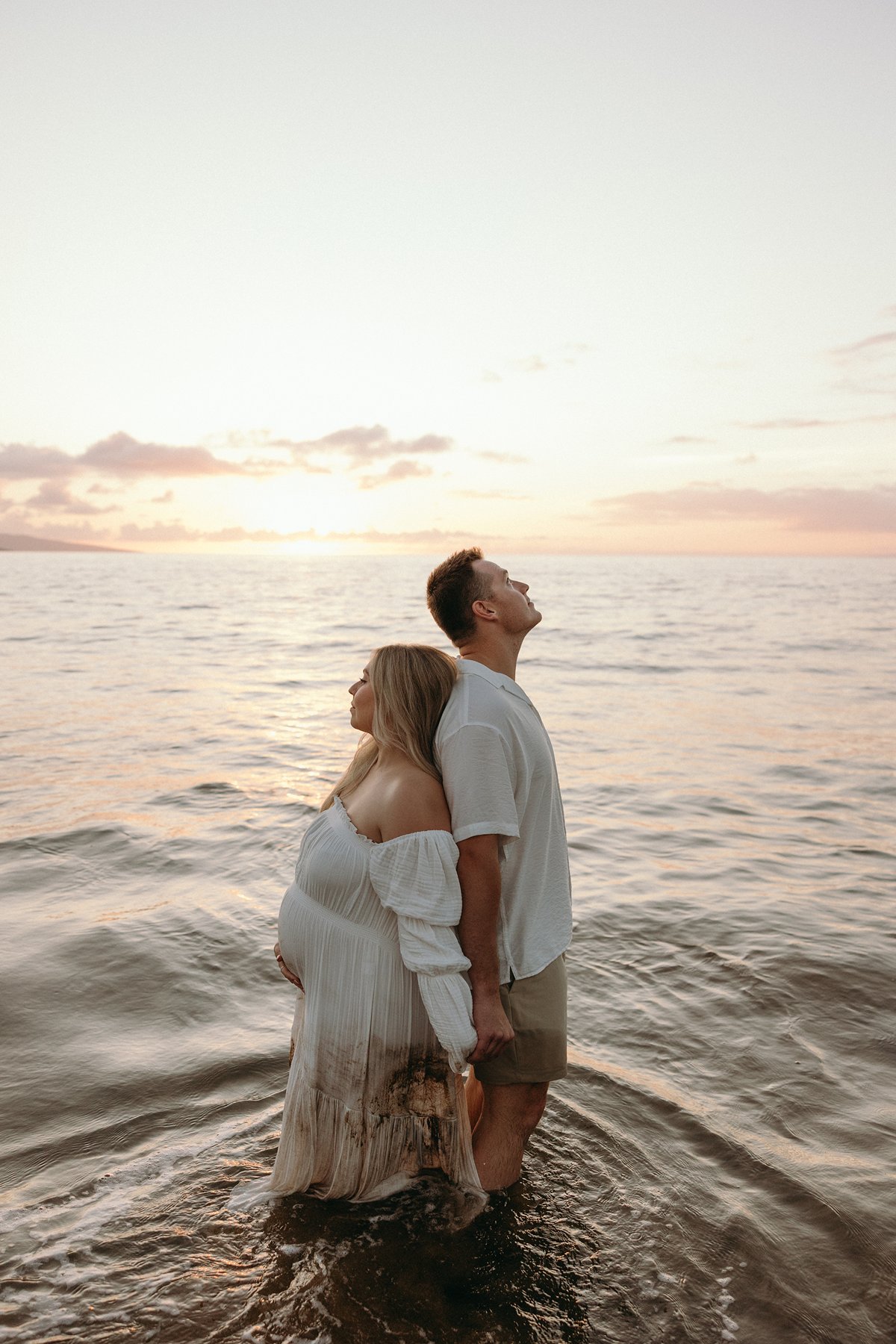 Pregnant couple standing in the ocean together at sunset during their Maui babymoon.