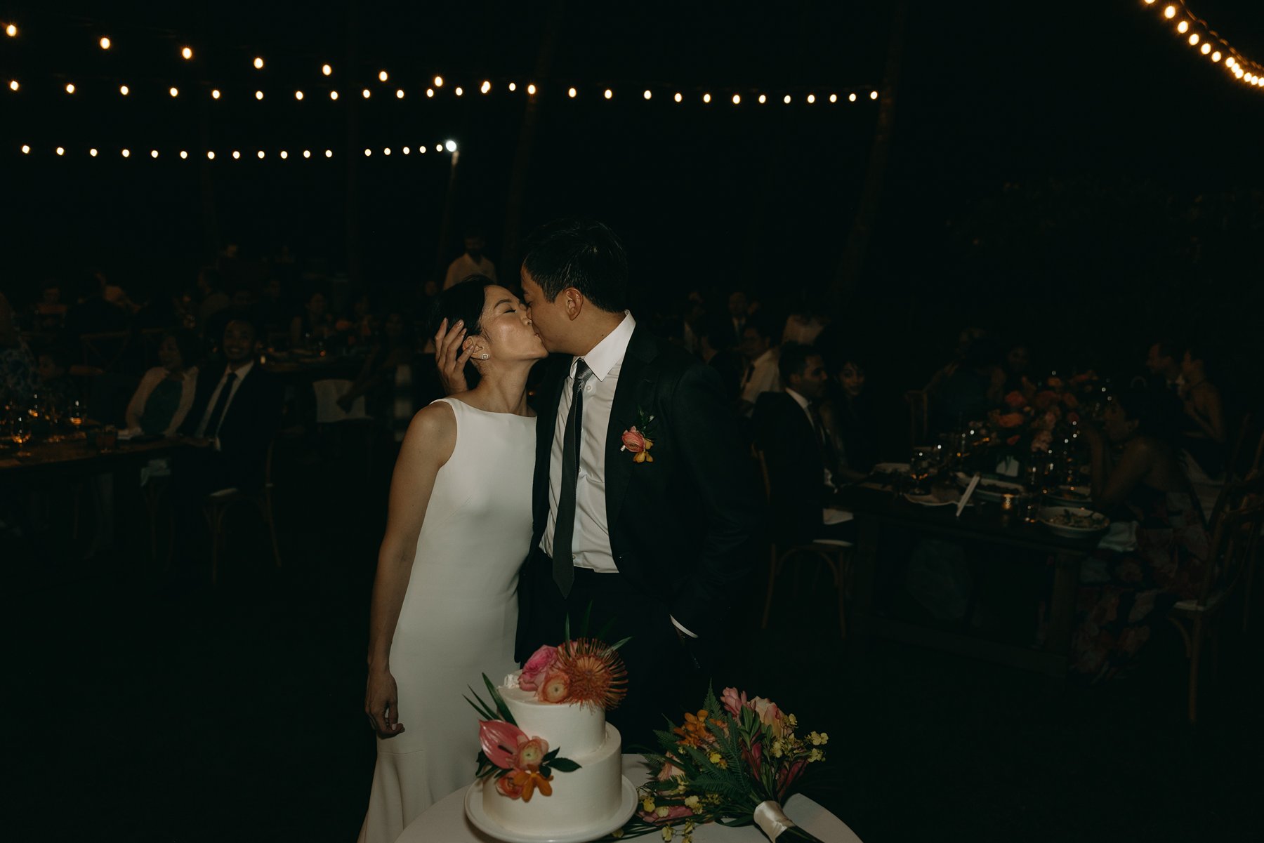 Bride and groom kissing at nighttime reception under string lights with wedding cake