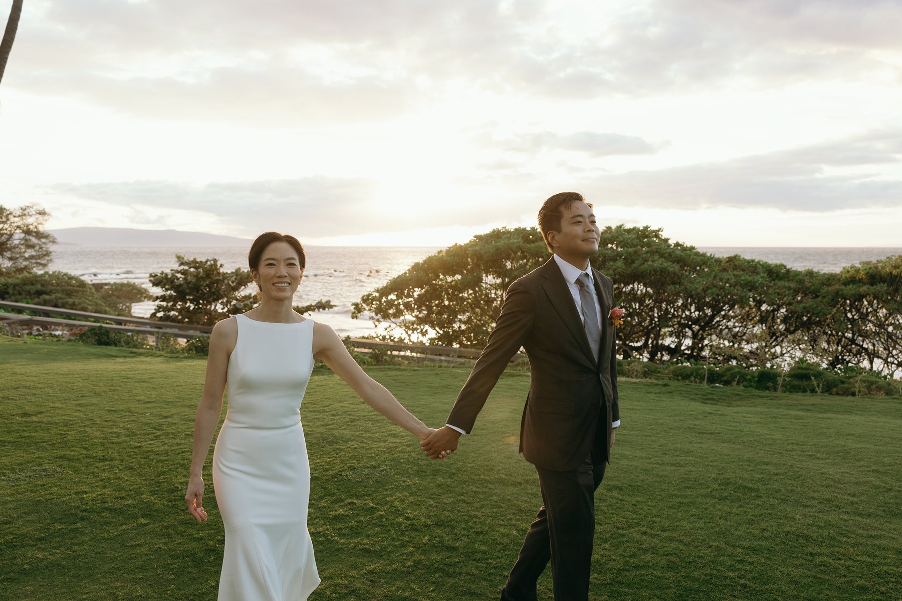 Bride and groom holding hands and walking on oceanfront lawn at sunset in Maui