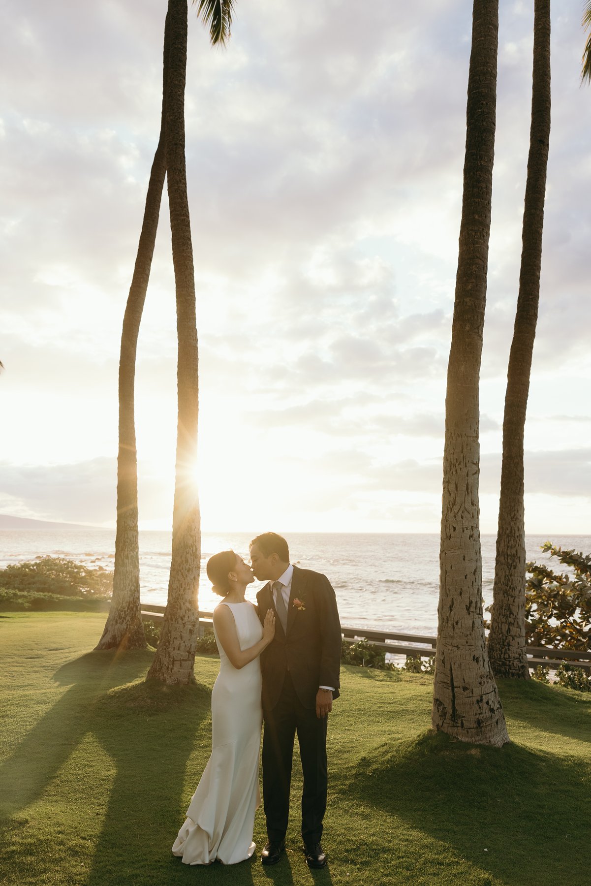 Bride and groom kissing under palm trees at sunset overlooking ocean in Hawaii