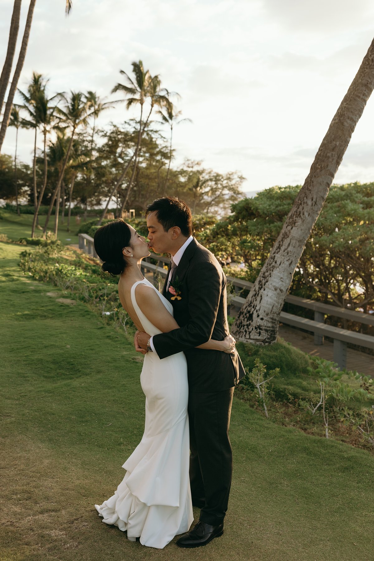 Bride and groom kissing on oceanfront lawn at sunset in Maui with palm trees and golden light