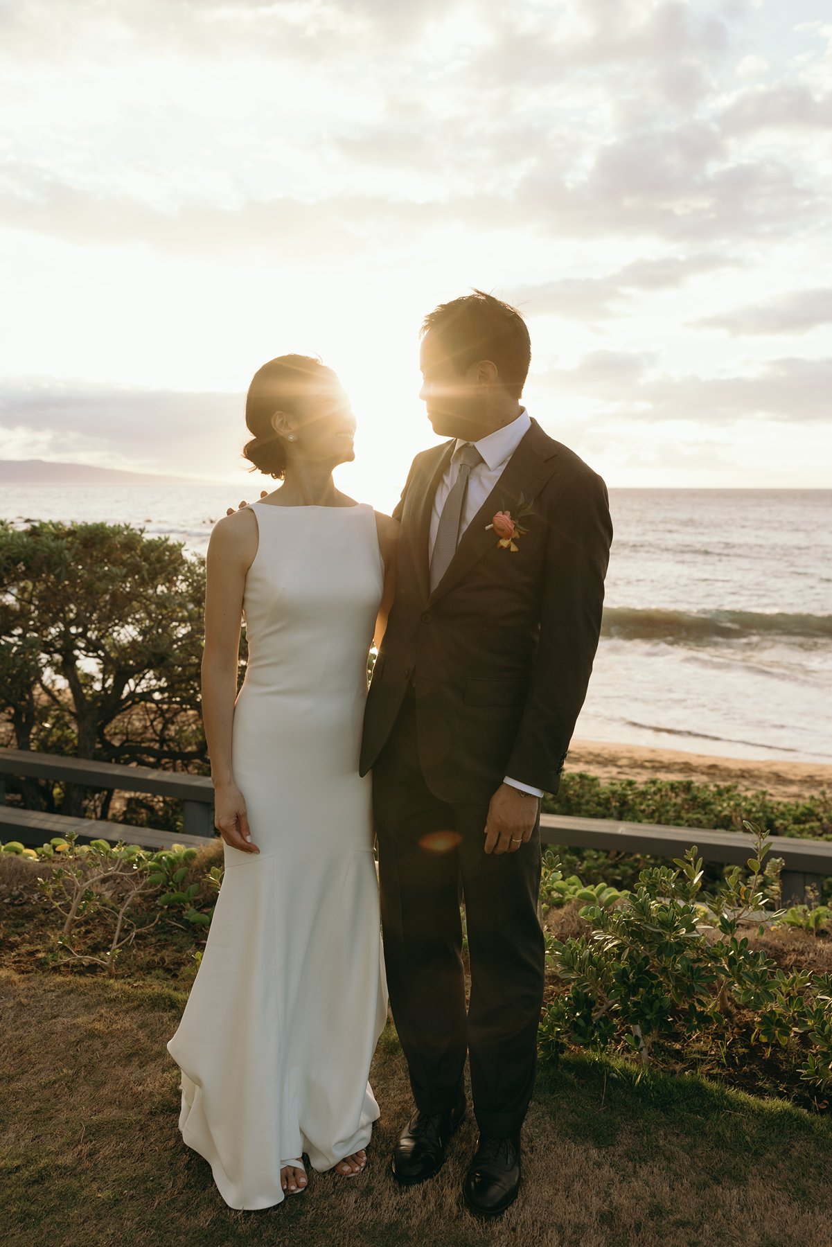 Bride and groom standing together with sun flare over ocean during Maui beach wedding portraits