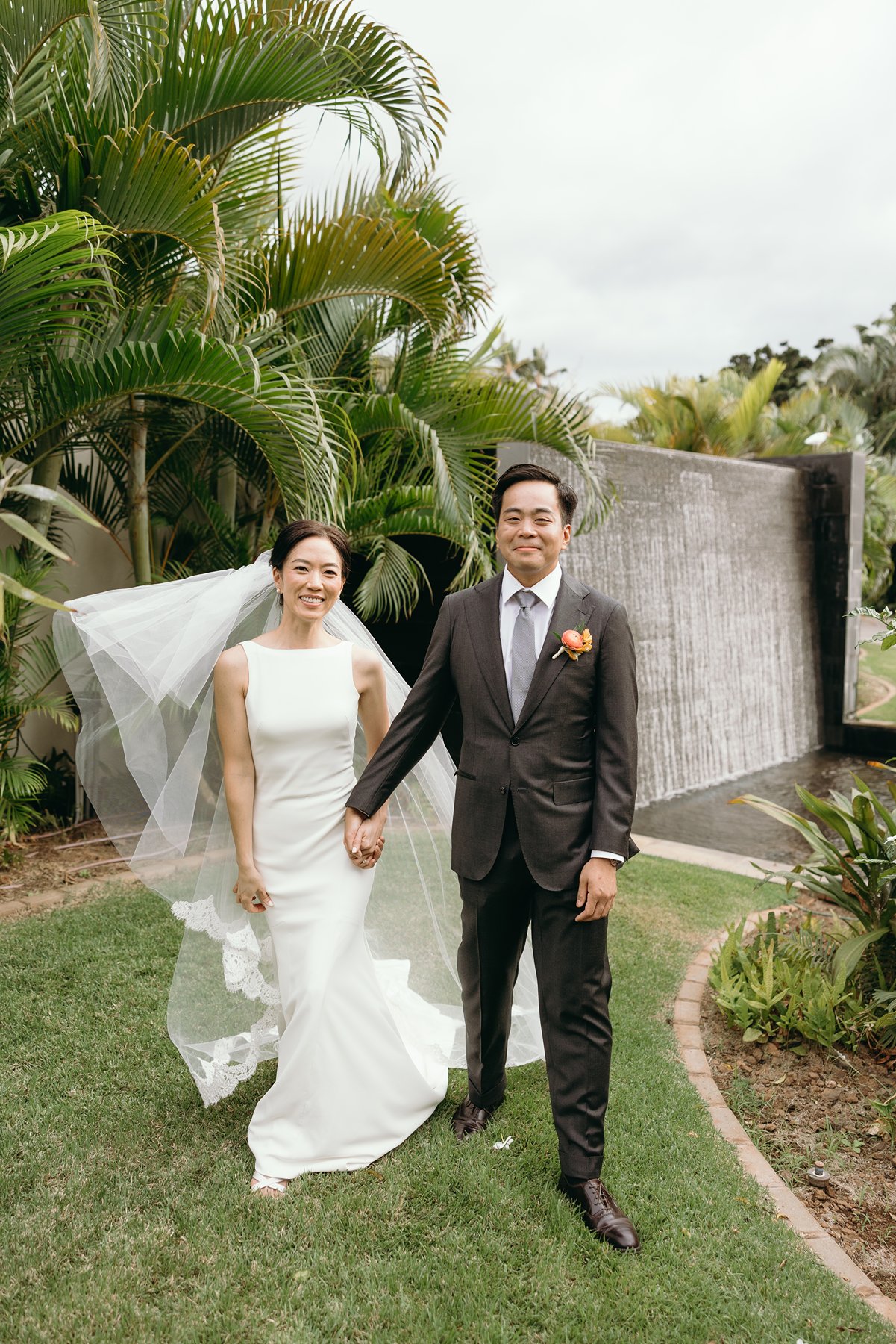 Bride and groom holding hands in tropical garden with waterfall at Maui resort wedding venue