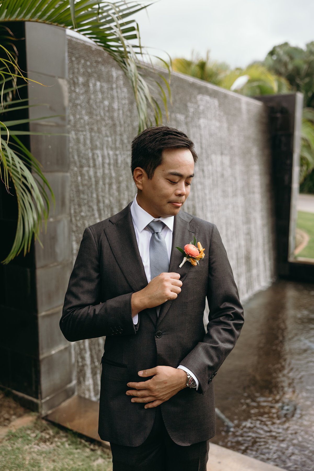 Groom adjusting boutonniere in front of waterfall at tropical resort wedding venue